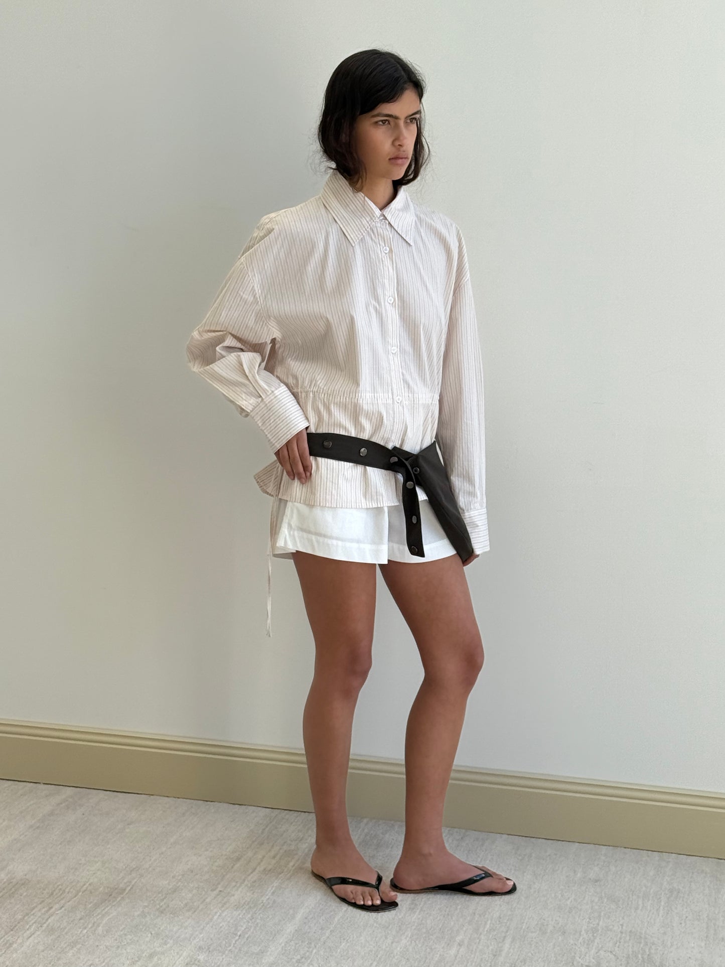 A woman stands indoors against a plain wall, wearing a loose, light-colored button-up shirt, white shorts, black flip-flops, and the Press Stud Saddle Bag - Dark Chocolate slung across her hips.