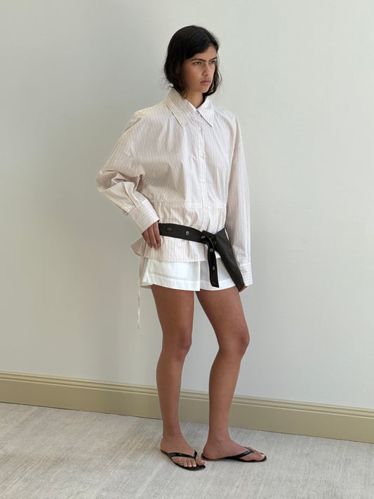 A woman stands indoors against a plain wall, wearing a loose, light-colored button-up shirt, white shorts, black flip-flops, and the Press Stud Saddle Bag - Dark Chocolate slung across her hips.