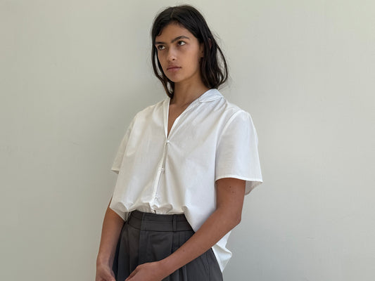 A person with long dark hair wears the Deiji Studios Sails Top in white and high-waisted gray trousers, standing against a plain light-colored wall with a neutral expression.