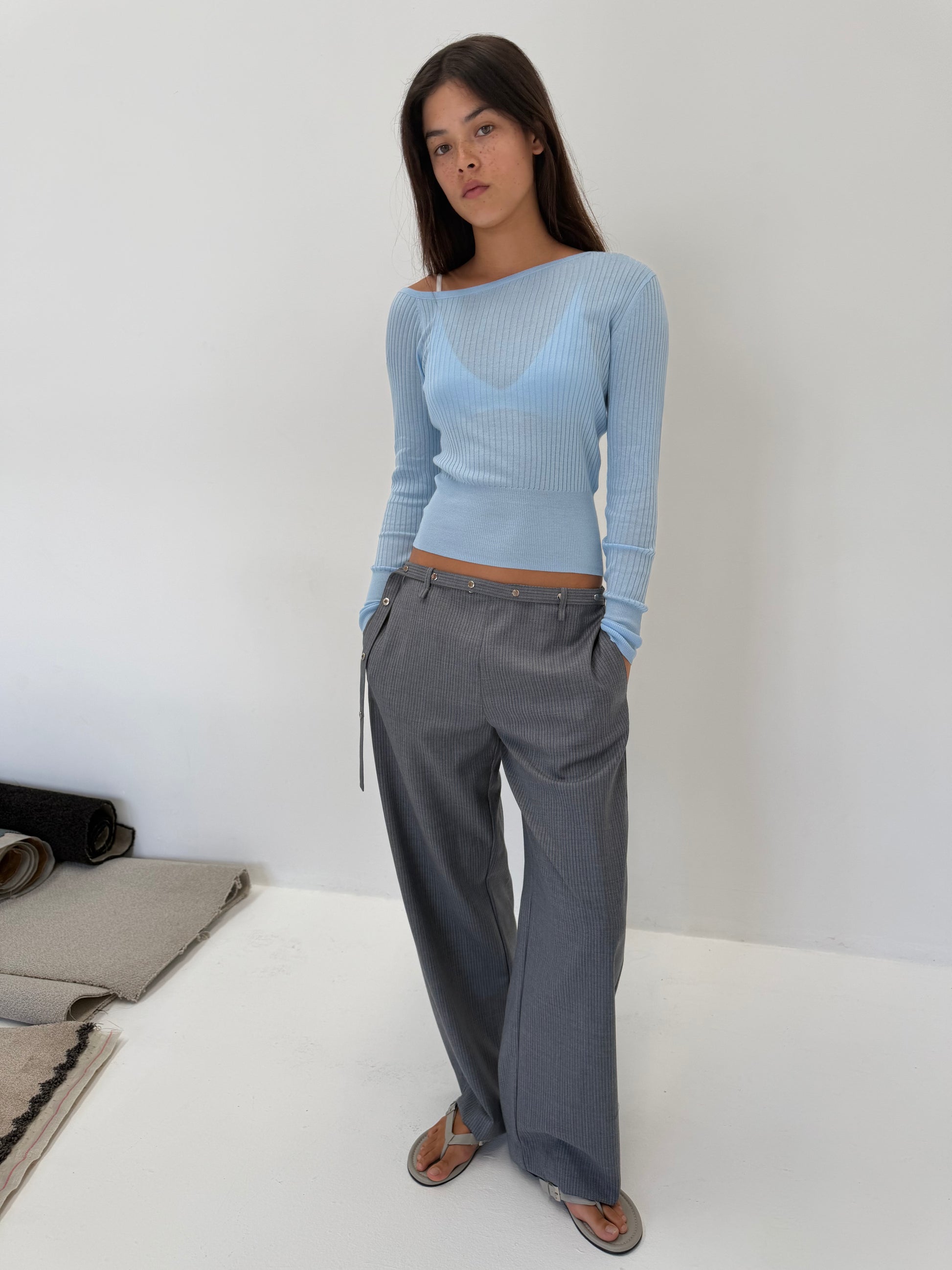 A woman wearing the Shoulder Knit Top - Blue over a blue bralette, paired with grey wide-leg pinstripe trousers with side ties and grey sandals.