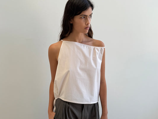 A woman with long dark hair wears the Simple Top - Dusty Stripe by Deiji Studios, a white one-shoulder sleeveless organic cotton top, paired with dark pants. She stands against a plain light background, gazing thoughtfully to the side.