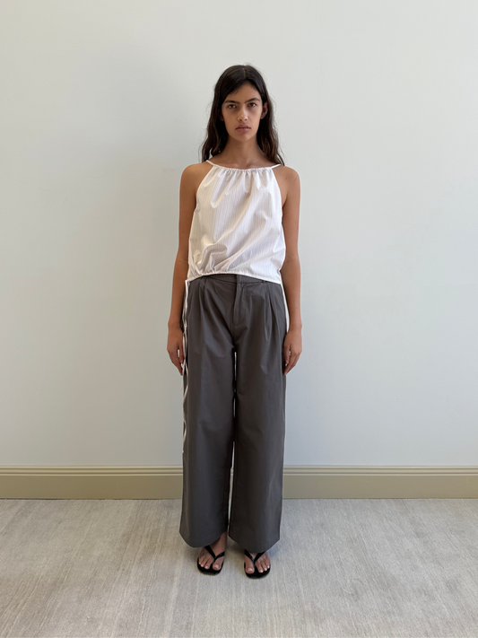 A woman with long dark hair stands against a plain light wall, wearing the Simple Top - Dusty Stripe by Deiji Studios, loose gray wide-leg pants, and black sandals. She has a neutral expression.