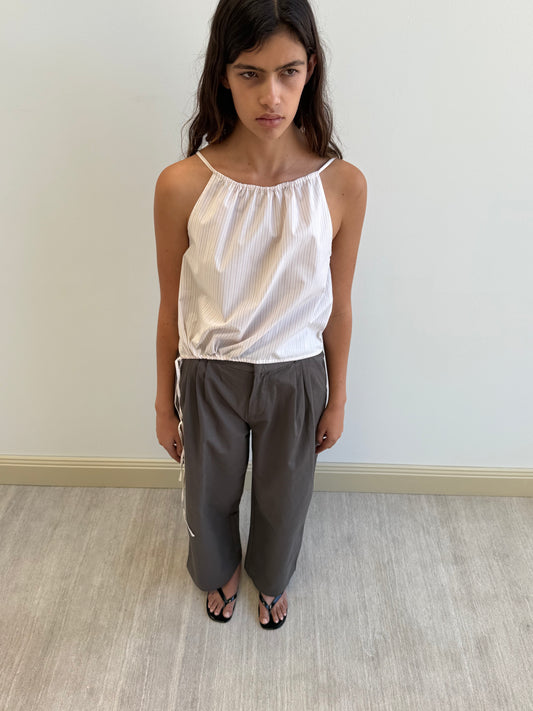A woman with long brown hair stands indoors against a white wall, wearing the Simple Top - Dusty Stripe by Deiji Studios, loose gray pants, and black sandals. She gazes slightly downward with a neutral expression.