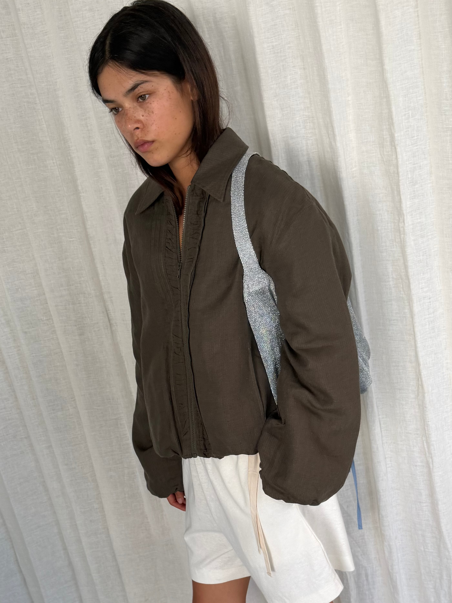 A young person with straight dark hair wears the Pintuck Jacket - Khaki Stripe, white shorts, and a silver backpack, standing in front of white curtains and looking slightly downward.