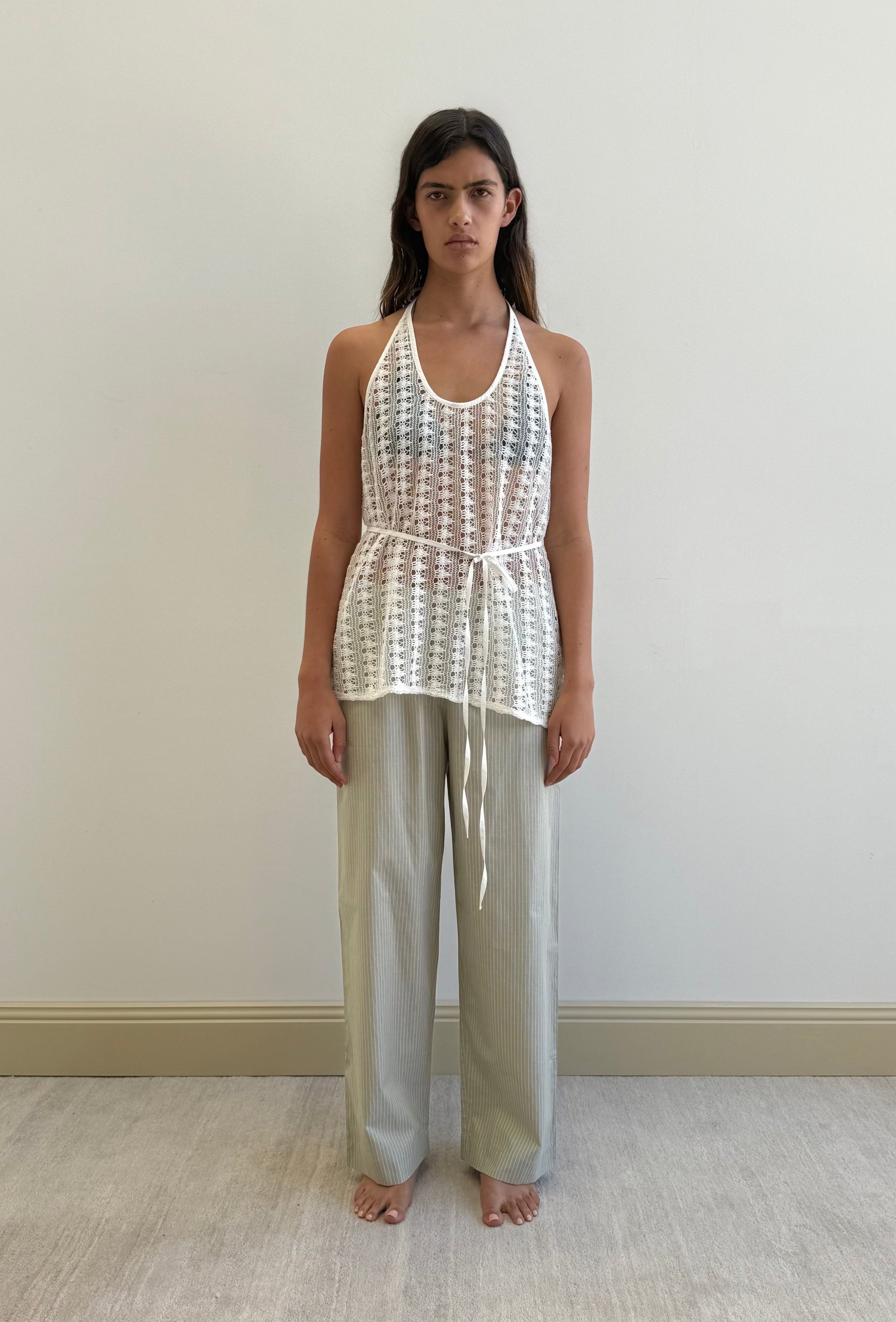 A woman stands barefoot against a plain wall, wearing the Ease Trouser in Pistachio Stripe from Deiji Studios and a sleeveless white crocheted top with a waist tie, looking directly at the camera with a neutral expression.