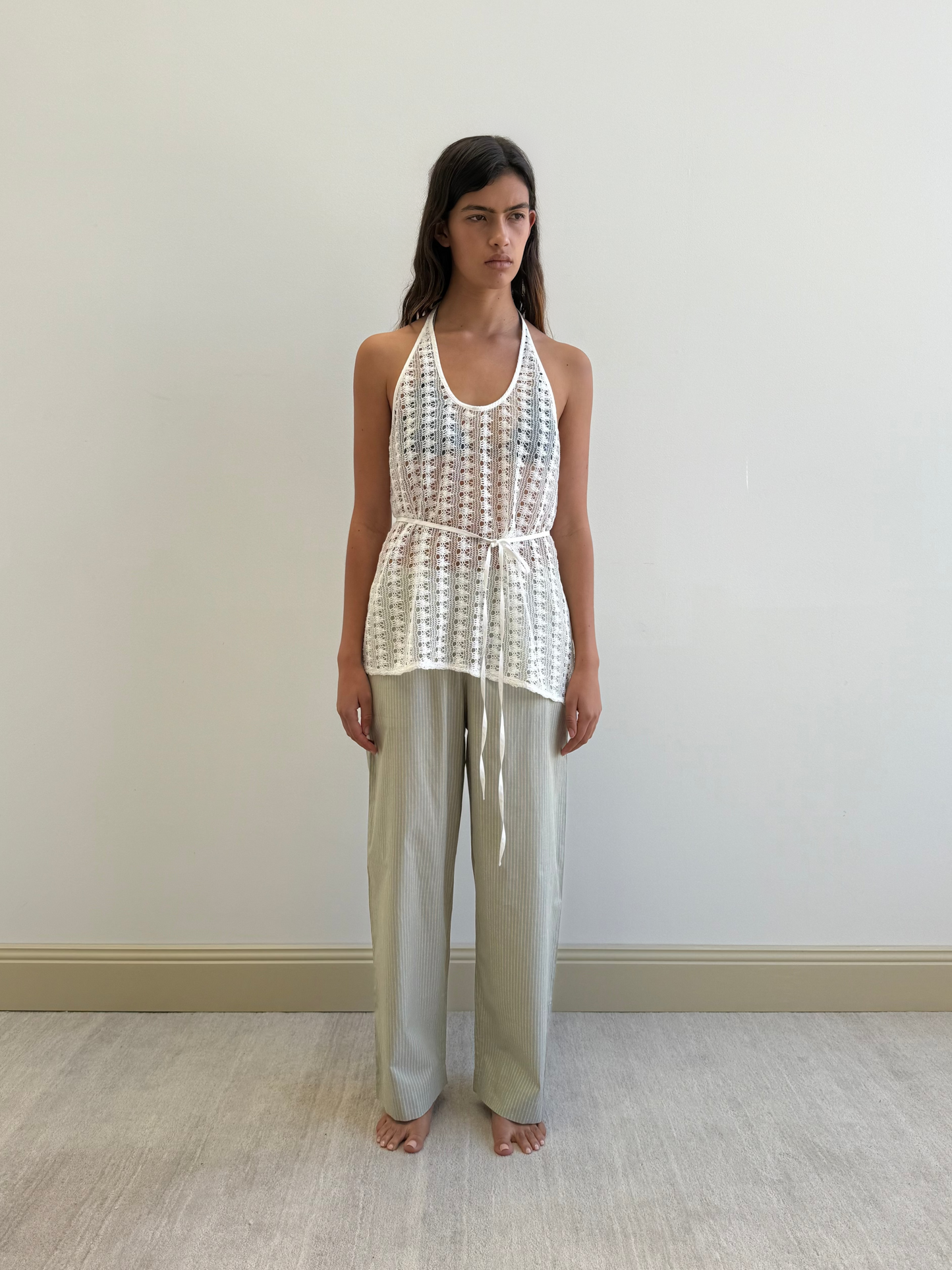 A woman stands barefoot indoors against a light wall, wearing a white sleeveless sheer crochet top with a tie belt over Ease Trouser - Pistachio Stripe by Deiji Studios. Her arms rest at her sides as she gazes forward.