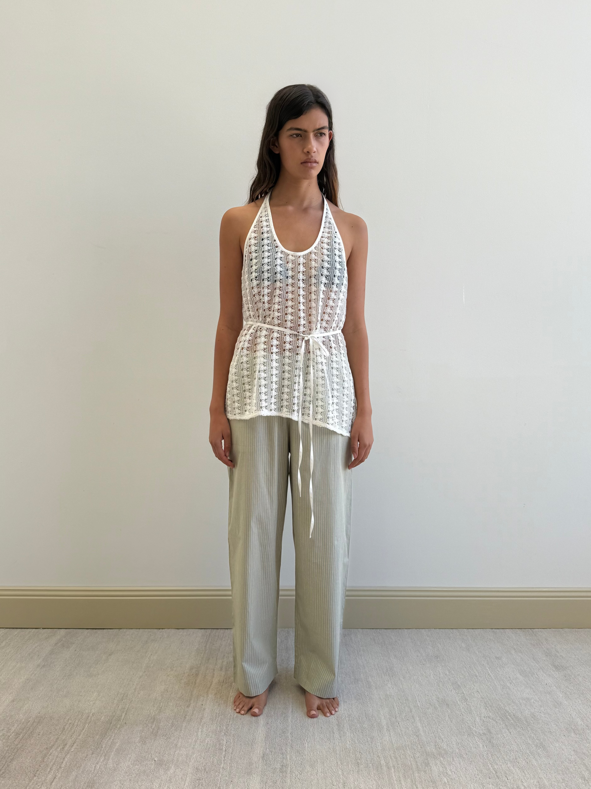 A woman stands barefoot indoors against a light wall, wearing a white sleeveless sheer crochet top with a tie belt over Ease Trouser - Pistachio Stripe by Deiji Studios. Her arms rest at her sides as she gazes forward.