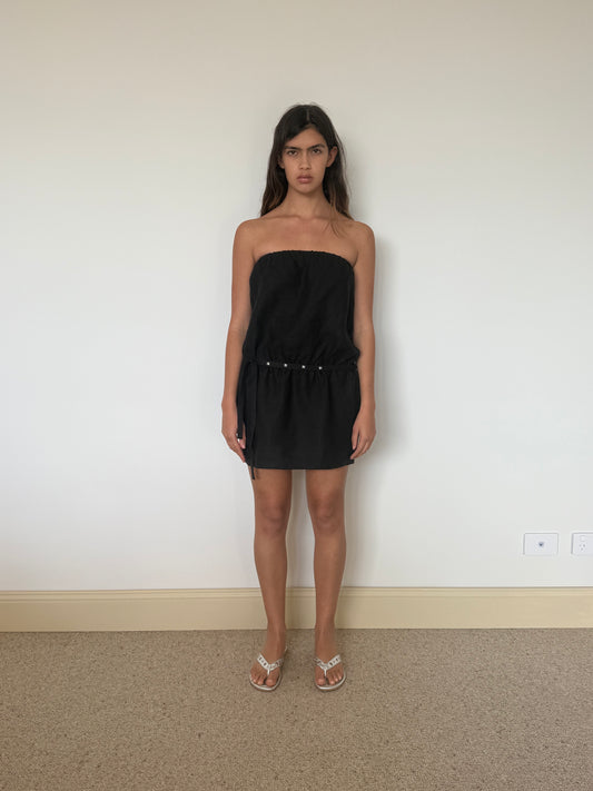 A young woman with long dark hair wears the Strapless Linen Dress - Black, paired with white sandals, standing indoors against a plain light-colored wall on a beige carpet.