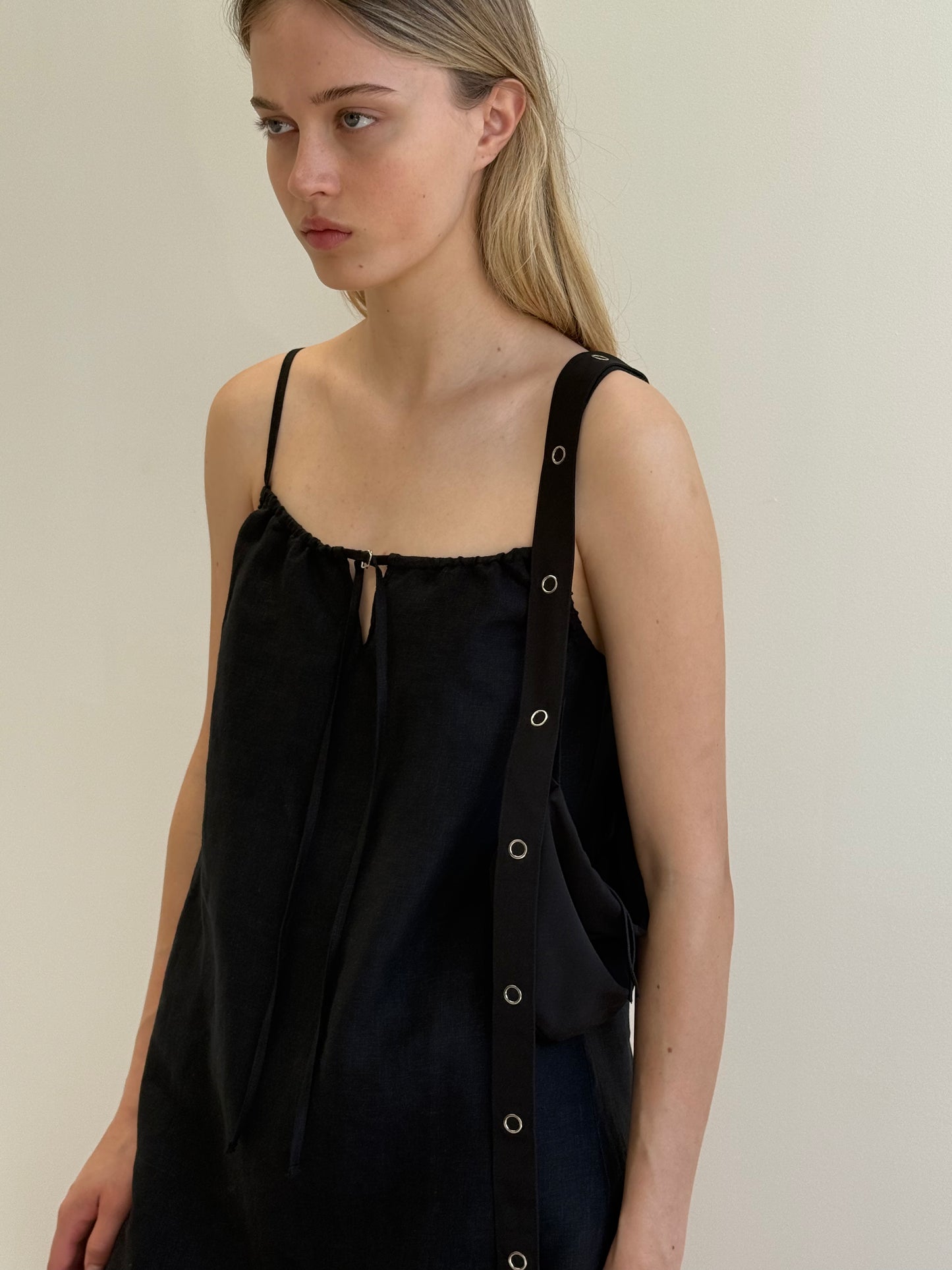 A woman with long blonde hair stands against a plain light background, wearing the Lettuce Mini Dress in black—a loose French linen dress with thin straps and grommet detailing over one shoulder.