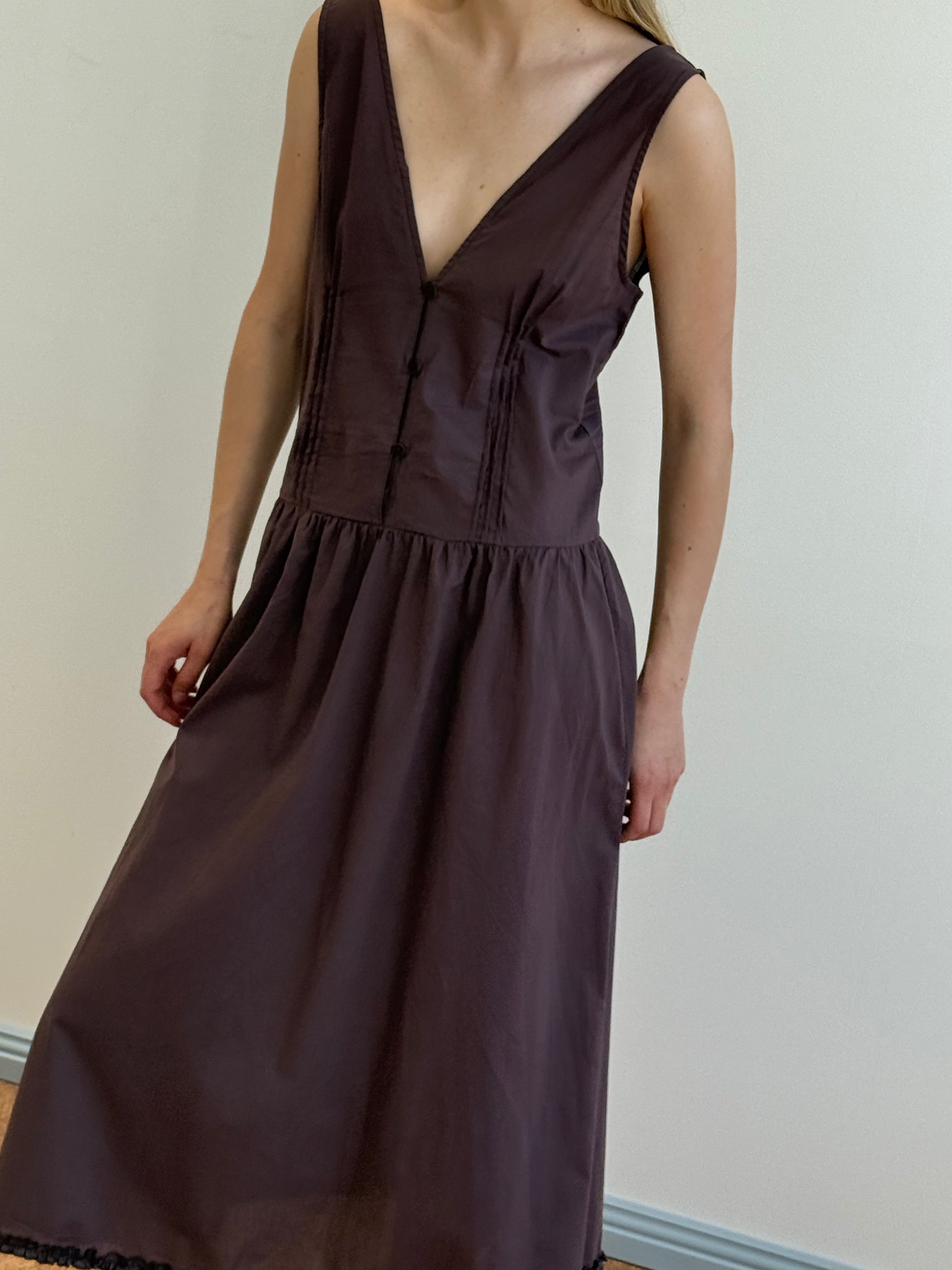 A woman wears the Pintuck Dress - Raisin by Deiji Studios, a sleeveless, deep V-neck dress with a fitted bodice and gathered skirt in dark brown organic cotton, standing before a plain light wall with her face partially out of frame.