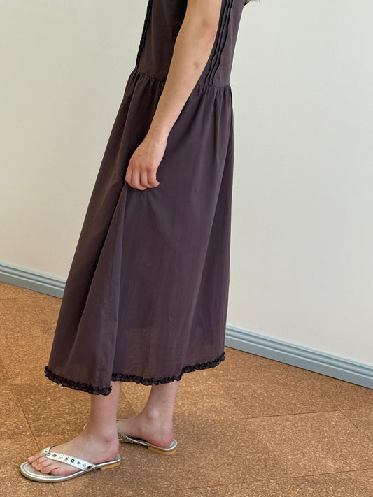 A person in a Pintuck Dress - Raisin, featuring short sleeves, organic cotton fabric, pintuck detail, lace-trimmed hem, and silver sandals stands on a brown floor by a light wall with blue baseboards.
