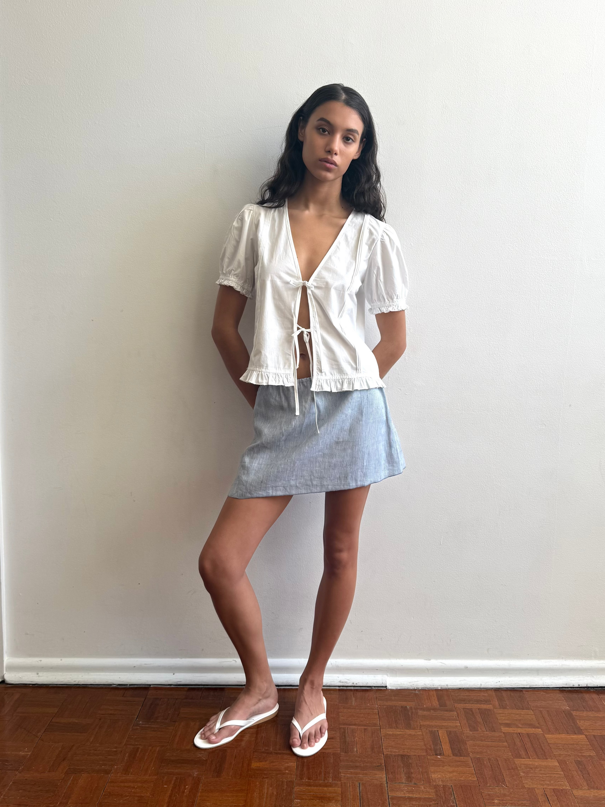 A woman stands indoors against a white wall, wearing a white short-sleeve blouse, the Babylock Skirt - Blue Stripe by Deiji Studios, and white sandals. She crosses one leg over the other on a wooden floor.