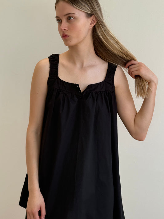 A woman with long blonde hair wears the Yoke Mini Dress - Black by Deiji Studios, a sleeveless, loose-fit mini dress, and looks to the side while holding her hair against a plain light background.