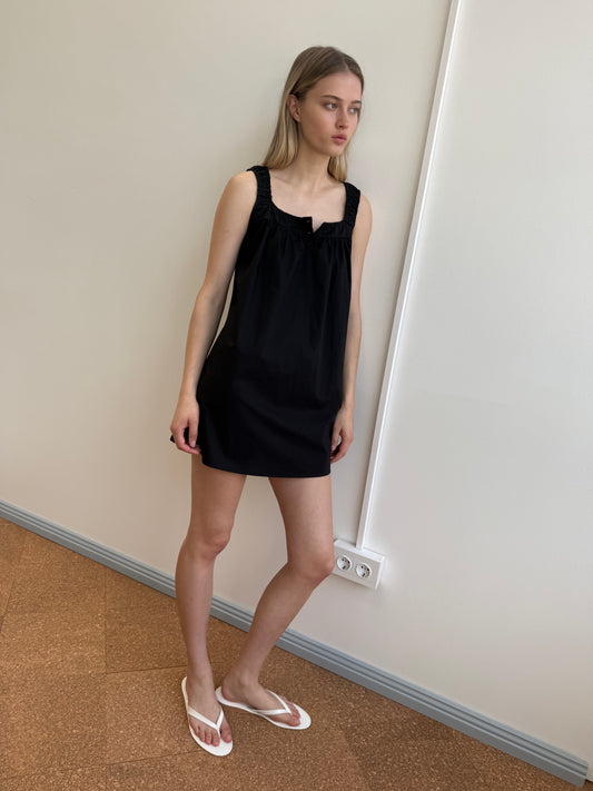 A woman stands indoors against a white wall, wearing the Yoke Mini Dress - Black from Deiji Studios and white flip-flops. She has straight light brown hair, a neutral expression, and one leg slightly bent.