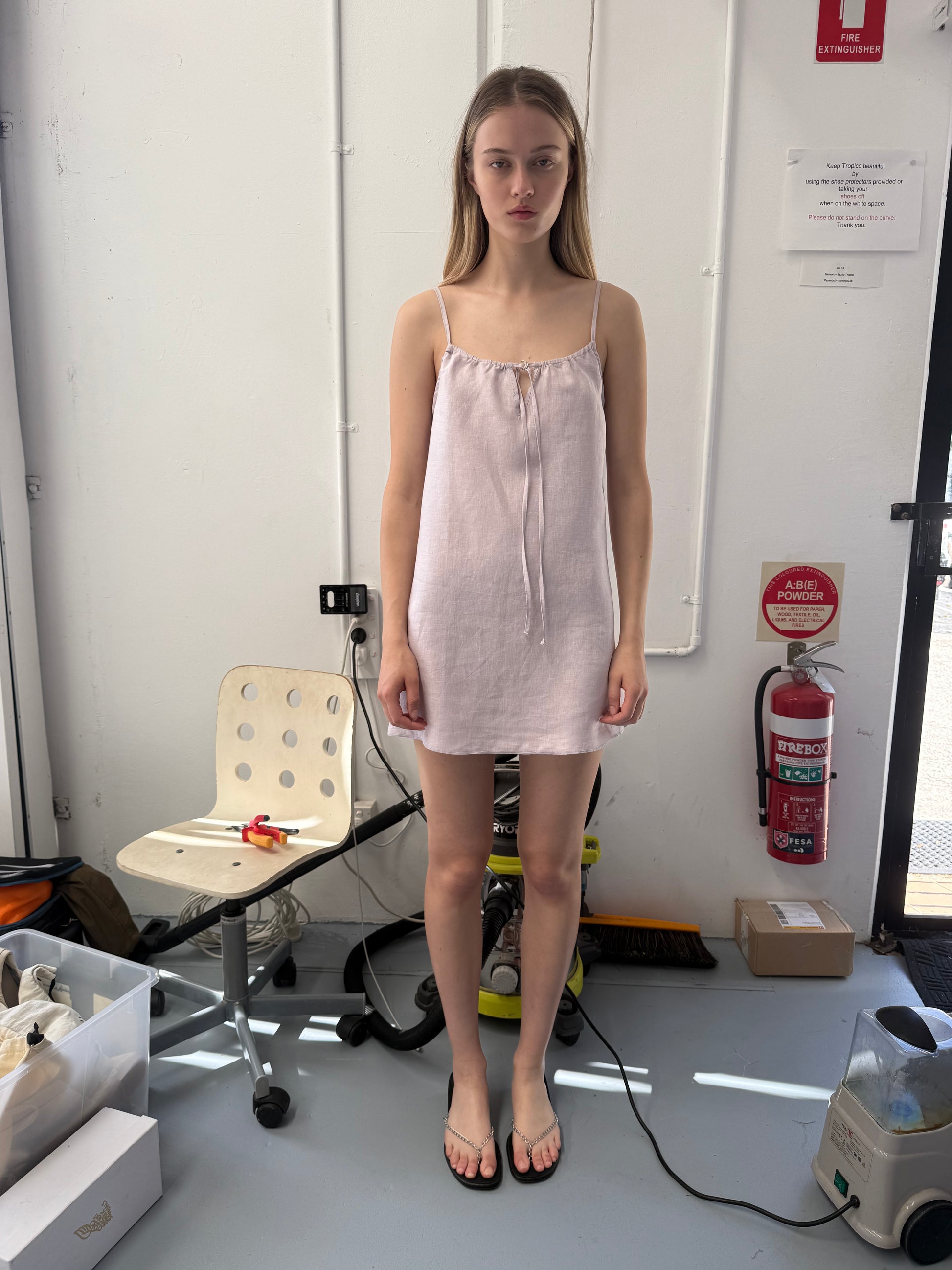 A young woman with long hair stands indoors wearing the Lettuce Mini Dress - Lilac and black sandals. Behind her are a white chair, tools, a fire extinguisher, and various items scattered on the floor against a white wall.