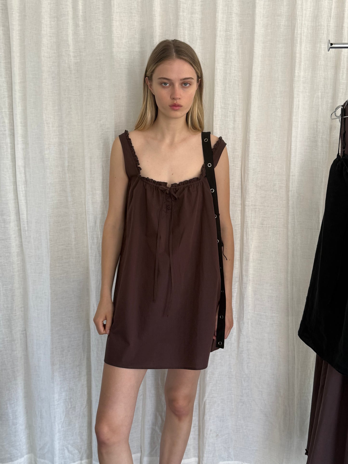 A young woman stands before white curtains wearing Deiji Studios' Button Frill Mini Dress in Umber, featuring ruffled straps and a tie chest. She carries a black shoulder bag and has straight blonde hair with a neutral expression.