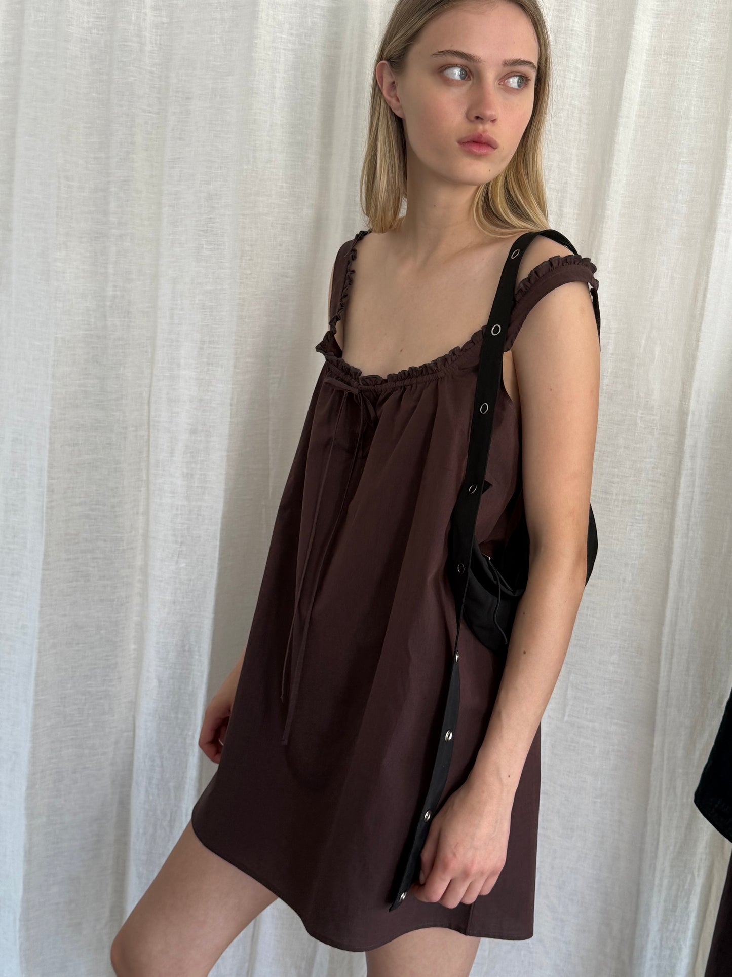 A young woman with long blonde hair wears the Button Frill Mini Dress in Umber and a black shoulder bag. She stands before a white curtain, looking to her right with a neutral expression.