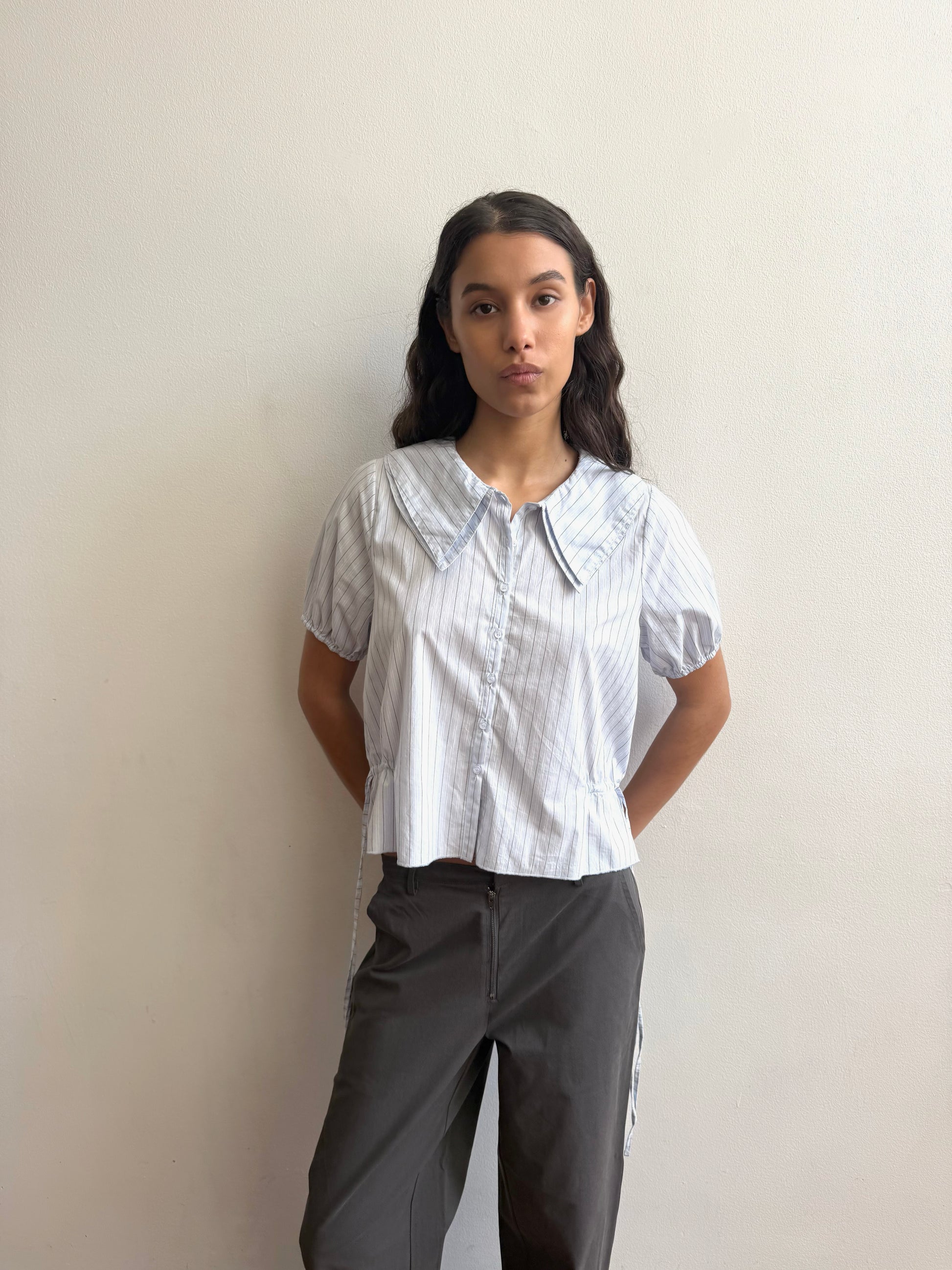 A young woman with long dark hair stands against a plain light wall, wearing Deiji Studios’ Double Collar Shirt – Mineral Stripe and dark gray pants, with her hands behind her back.