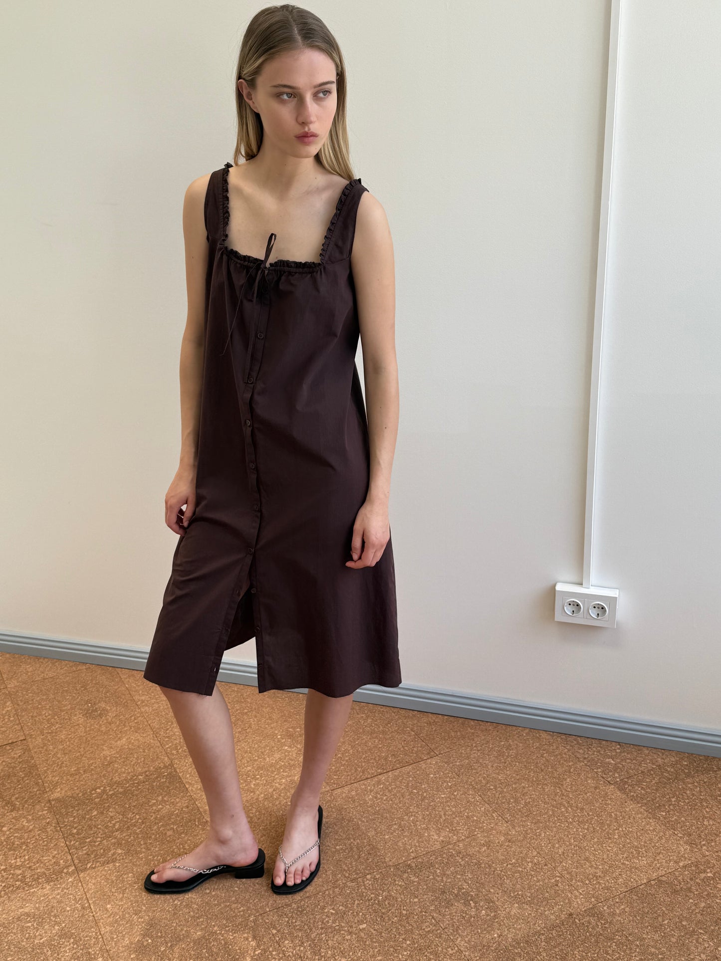 A woman with long blonde hair stands indoors, wearing the sleeveless, knee-length Button Sweep Dress in Umber made from organic cotton and black sandals. She faces slightly to the side with a neutral expression against a plain wall.