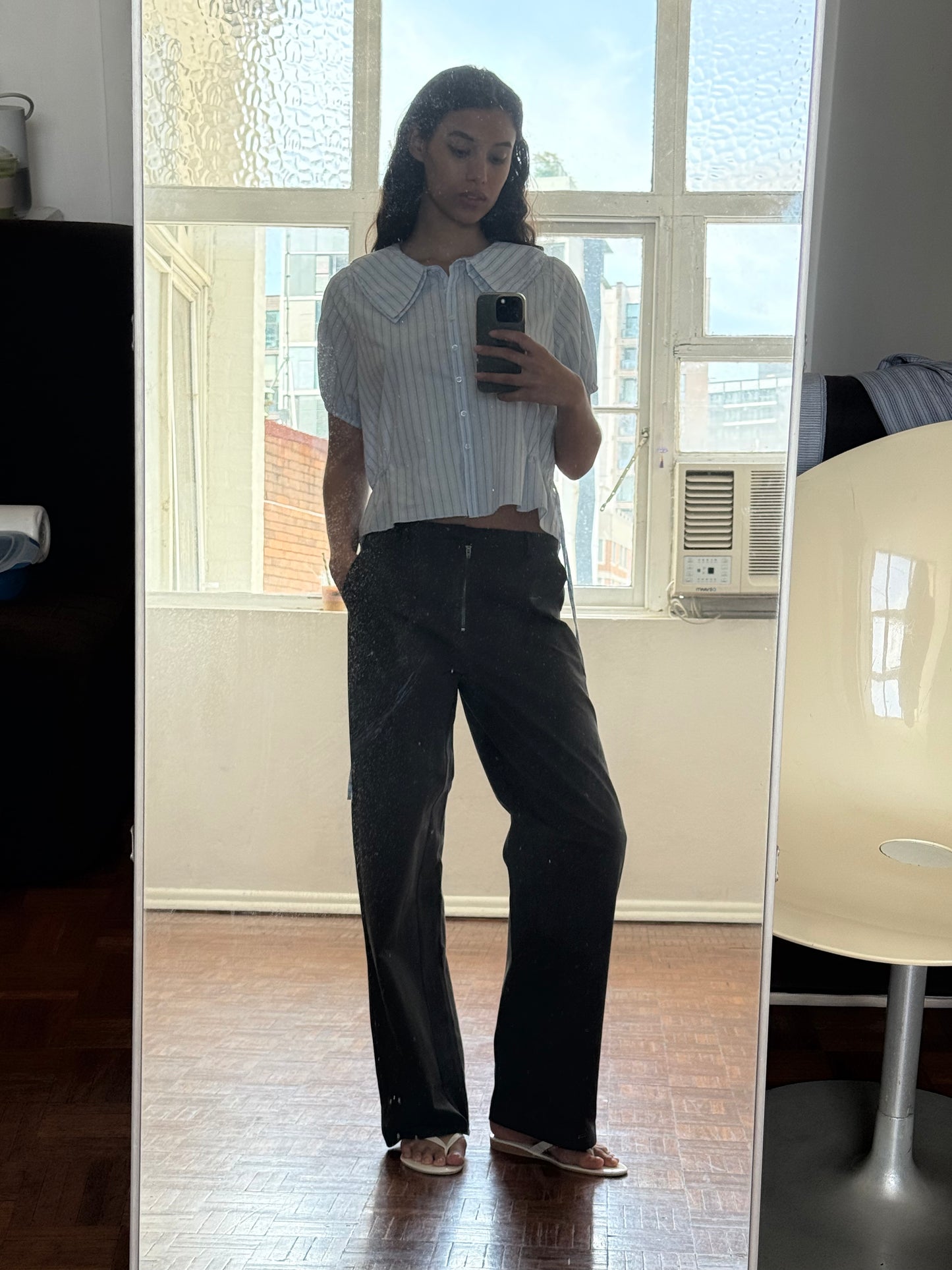 A person stands indoors taking a mirror selfie, wearing the Deiji Studios Double Collar Shirt - Mineral Stripe with black pants and white sandals. Behind them are a window, an air conditioner, and a white chair.