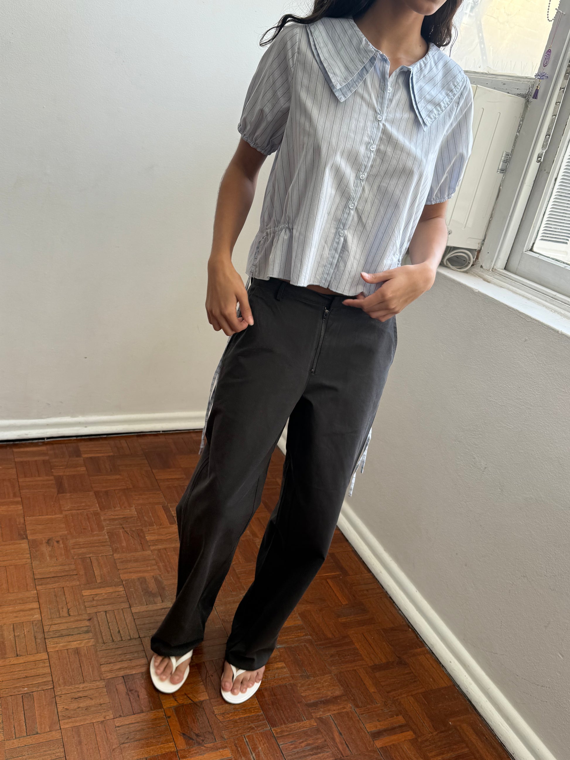 A person stands indoors on a wooden floor near a window, wearing black pants and the Double Collar Shirt - Mineral Stripe from Deiji Studios, paired with white sandals. Their head is partially out of the frame.