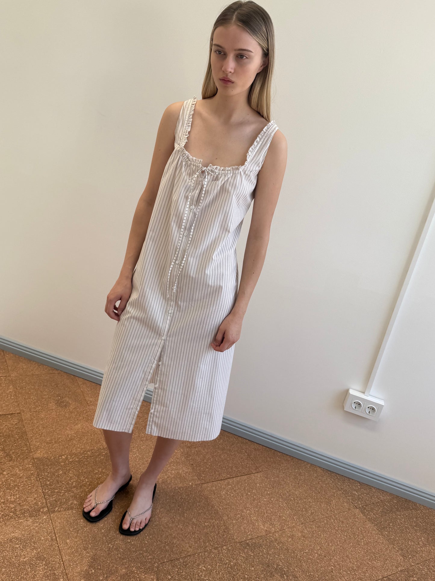 A young woman with straight, long hair stands indoors against a plain wall, wearing the Button Sweep Dress - Gull Stripe, a white sleeveless maxi dress with subtle vertical stripes, paired with black flip-flops and a neutral expression.