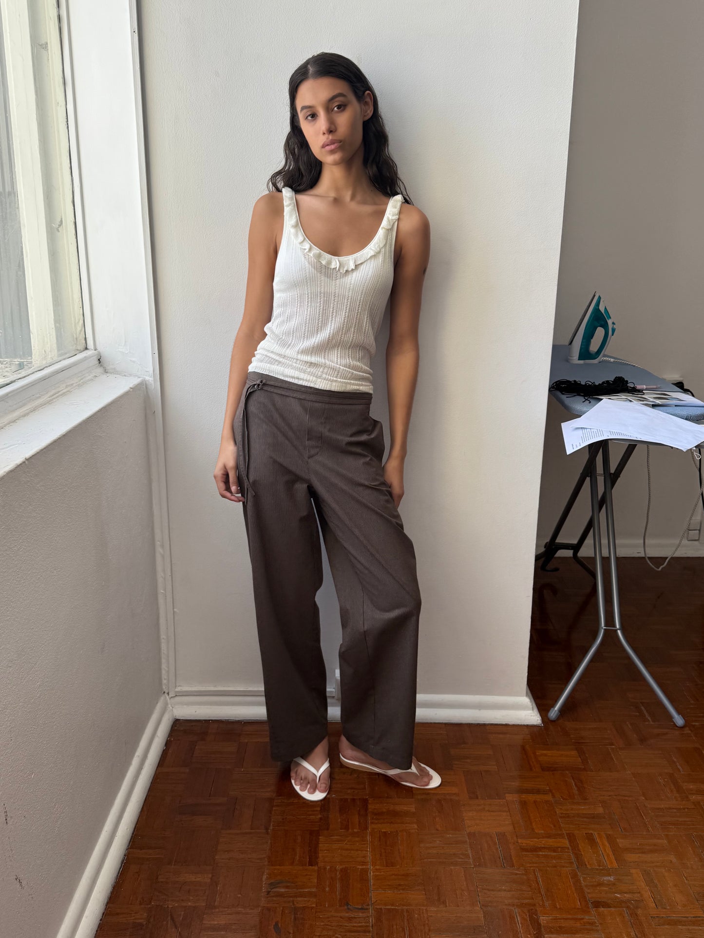 A woman in the Deiji Studios Neck Ruffle Knit - Off White and brown wide-leg pants stands by a white wall and window on a wooden floor, with an ironing board, iron, and papers in the background.