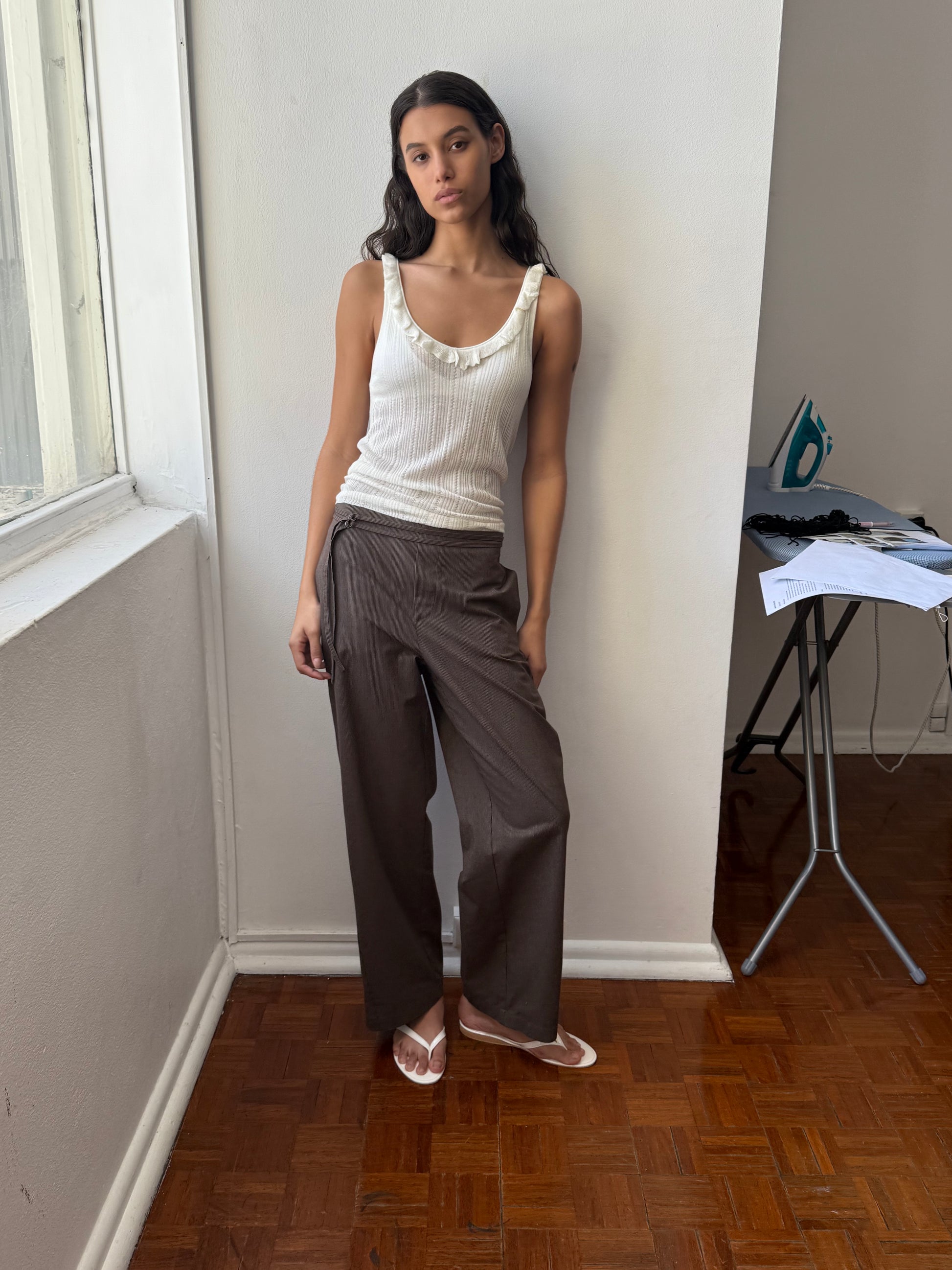 A woman in the Deiji Studios Neck Ruffle Knit - Off White and brown wide-leg pants stands by a white wall and window on a wooden floor, with an ironing board, iron, and papers in the background.