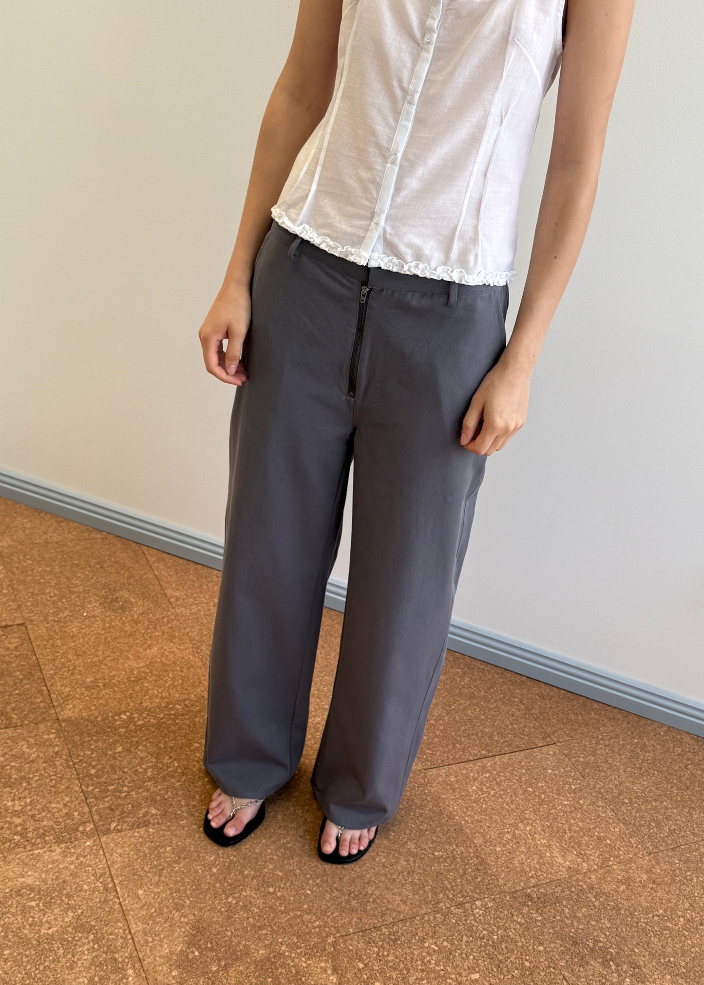 A person stands indoors wearing a sleeveless white blouse, Deiji Studios Zip Pant - Charcoal in organic cotton twill, and black sandals. The image is cropped at the neck and ankles, with a neutral wall and wood floor background.