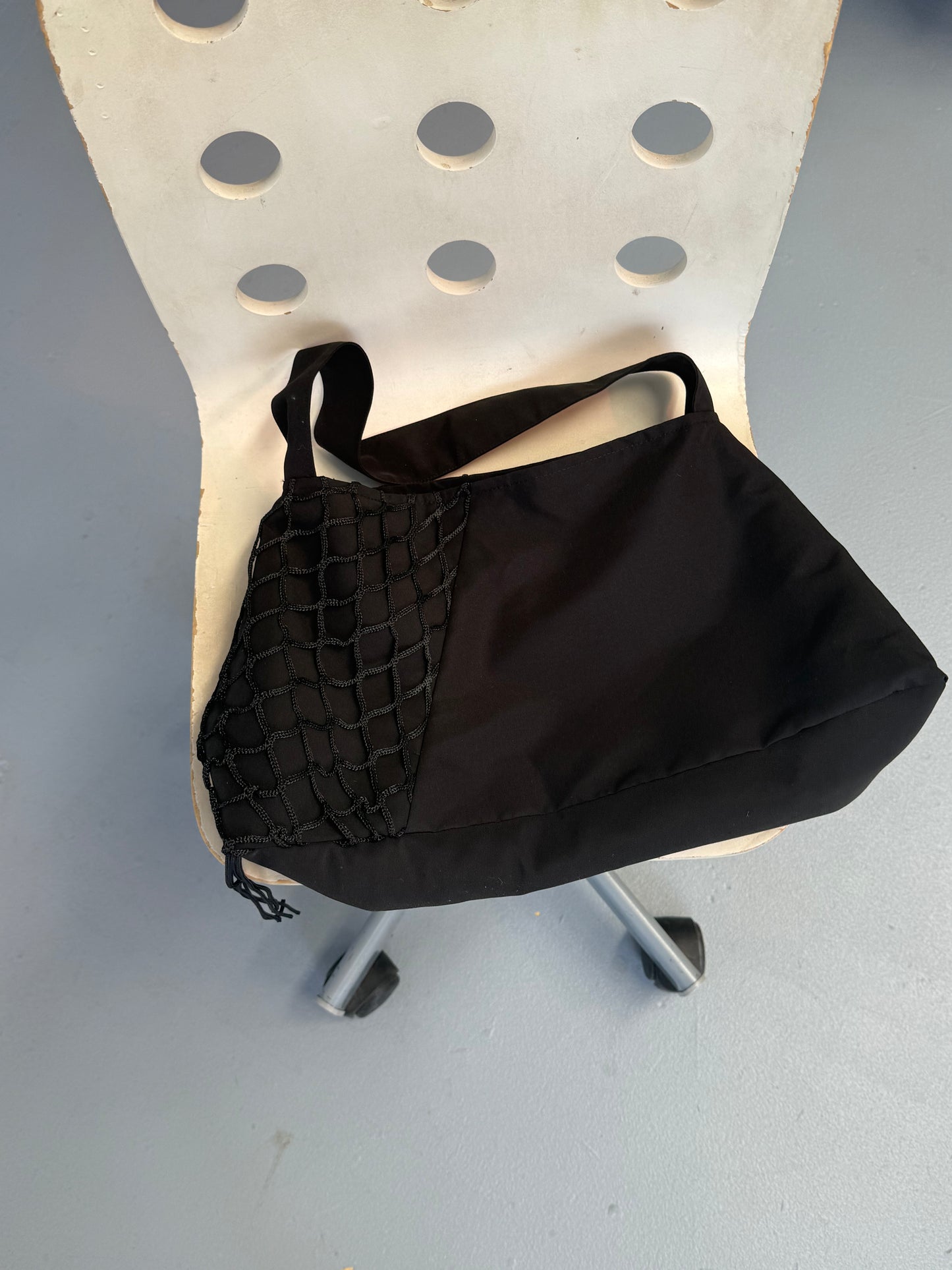 The Net Contrast Bag - Black from Deiji Studios, made with recycled polyester, sits on a white chair with circular cutouts, set against a light gray floor.