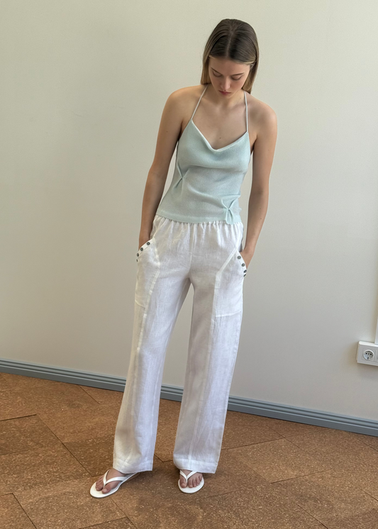 A woman stands indoors with her hands in the pockets of our Angle Pocket Pant - White, paired with a light blue halter top and white sandals. The clean look against a soft backdrop exudes effortless organic style.
