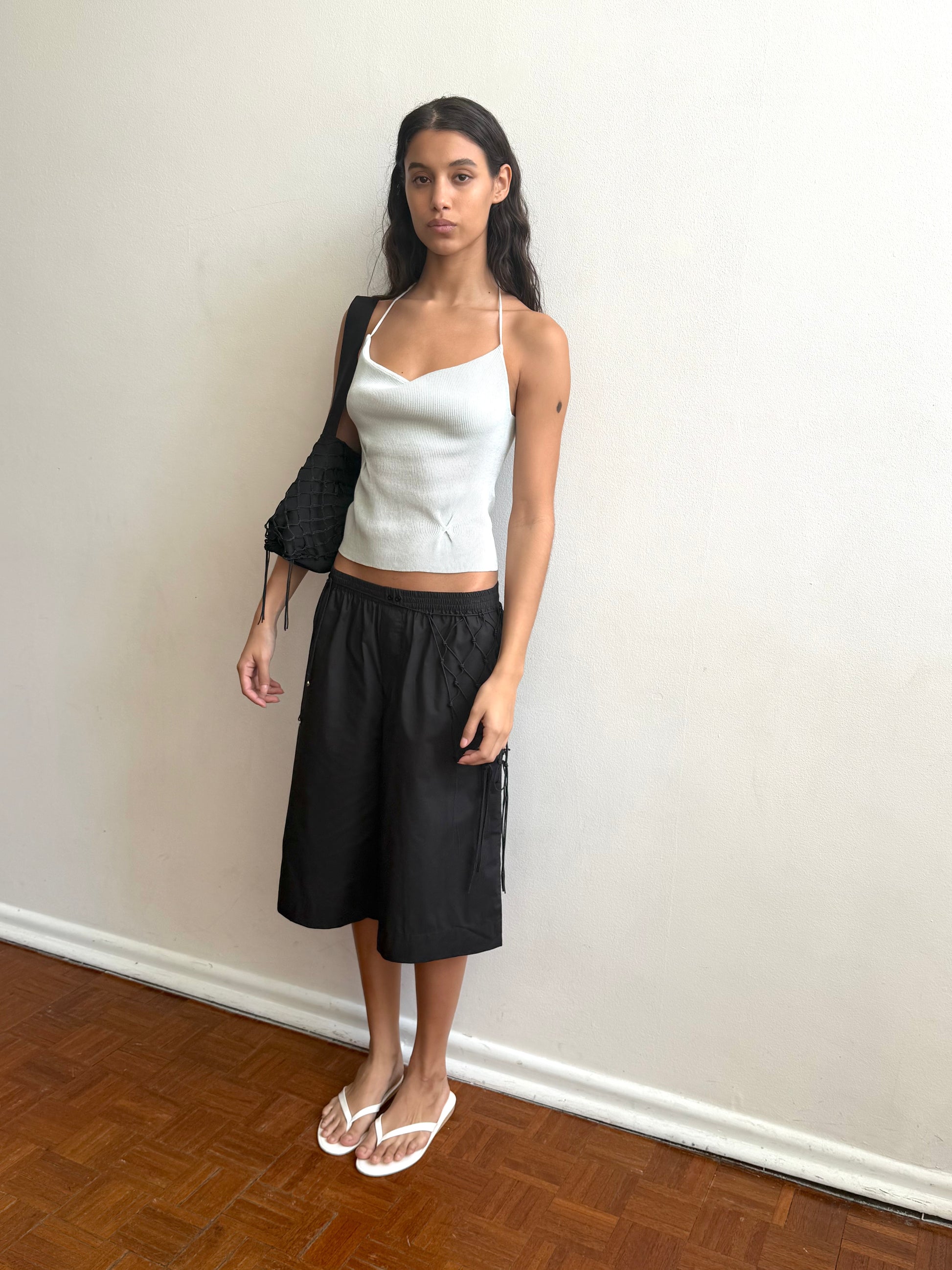 A woman with long dark hair and a neutral expression stands indoors against a plain white wall, wearing the Deiji Studios Fine Knot Tank in Ice Blue, a black knee-length skirt, white flip-flops, and holding a black purse.