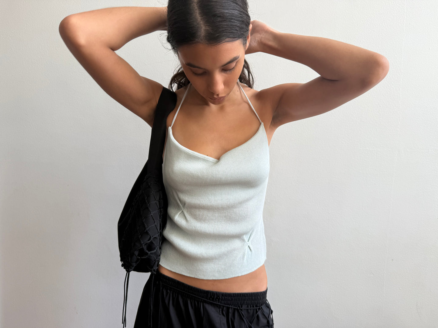 A woman with dark hair in a low ponytail adjusts her Deiji Studios Fine Knot Tank in Ice Blue while wearing black pants and a textured black shoulder bag, standing against a plain white background.