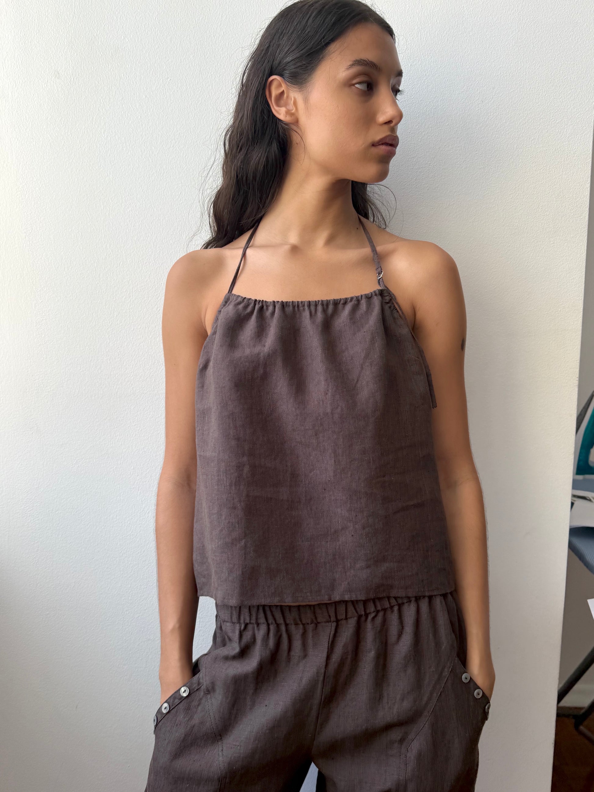 A woman with long dark hair stands against a light wall, wearing the O Ring Halter - Bark from Deiji Studios and matching pants, hands in her pockets, gazing to the side.
