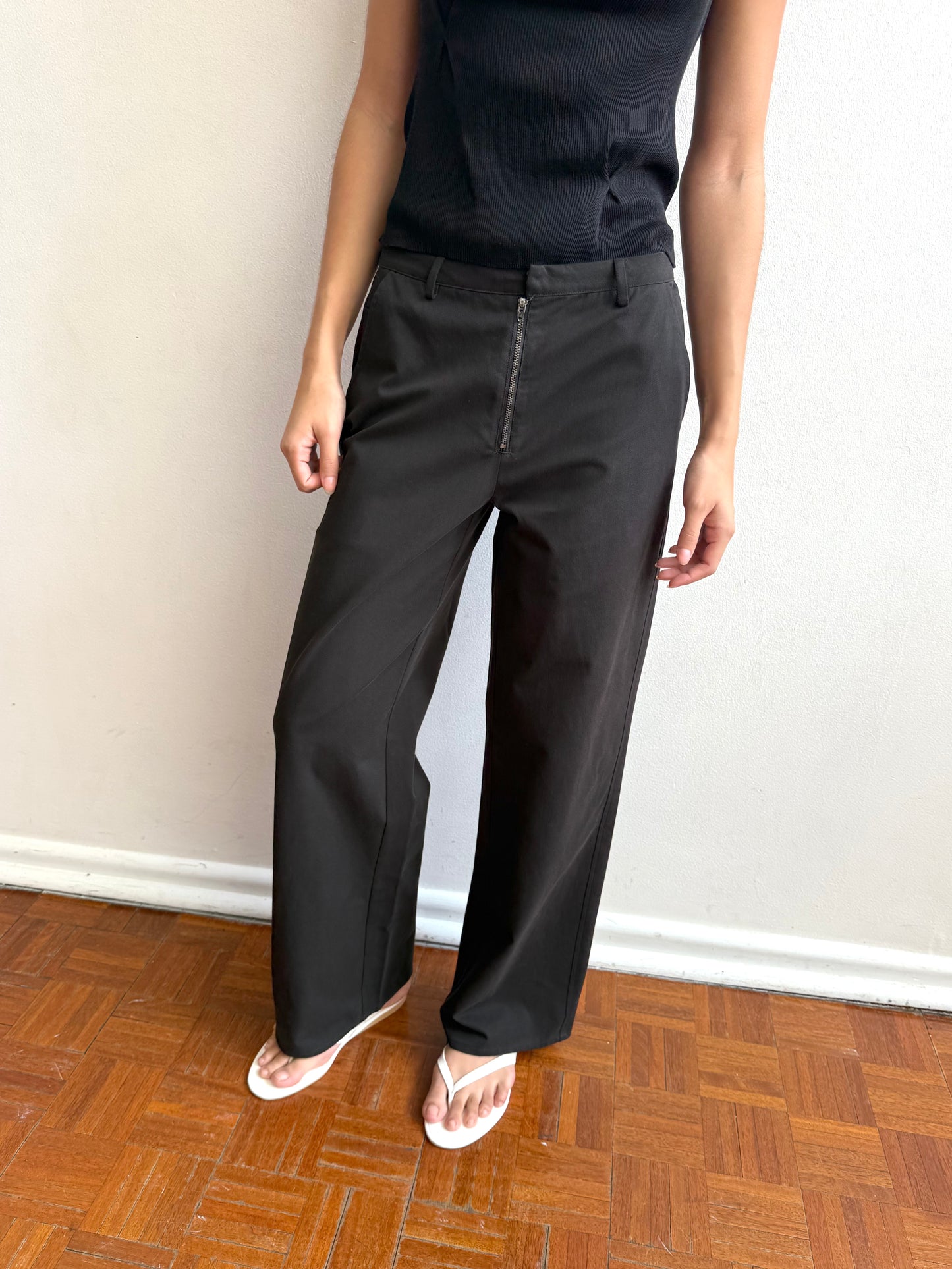 A person stands indoors against a white wall, wearing the Zip Pant - Black: high-waisted, wide-leg zip pants in organic cotton twill with a relaxed fit, paired with a sleeveless black top and white sandals. Only the torso and legs are visible.