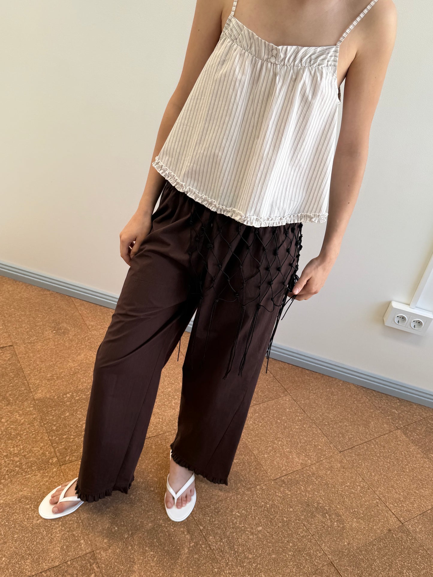 A person stands indoors on a light brown floor wearing the Yoke Top - Gull Stripe, a white pleated organic cotton spaghetti strap top, paired with loose brown pants featuring black netted fringe and white flip-flops.