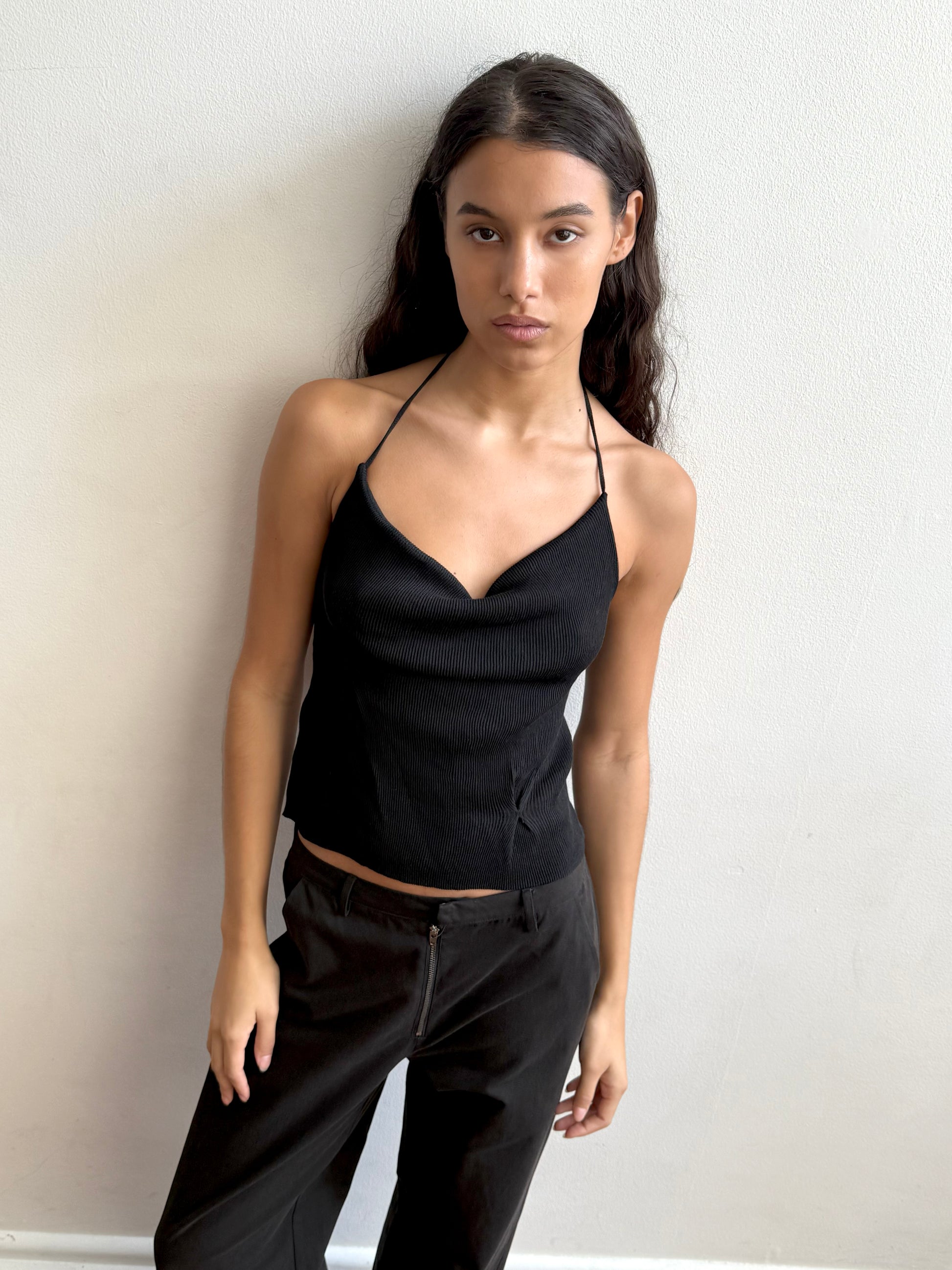 A woman with long dark hair models the Fine Knot Tank - Black and black pants, standing against a plain white wall and looking directly at the camera with a neutral expression.