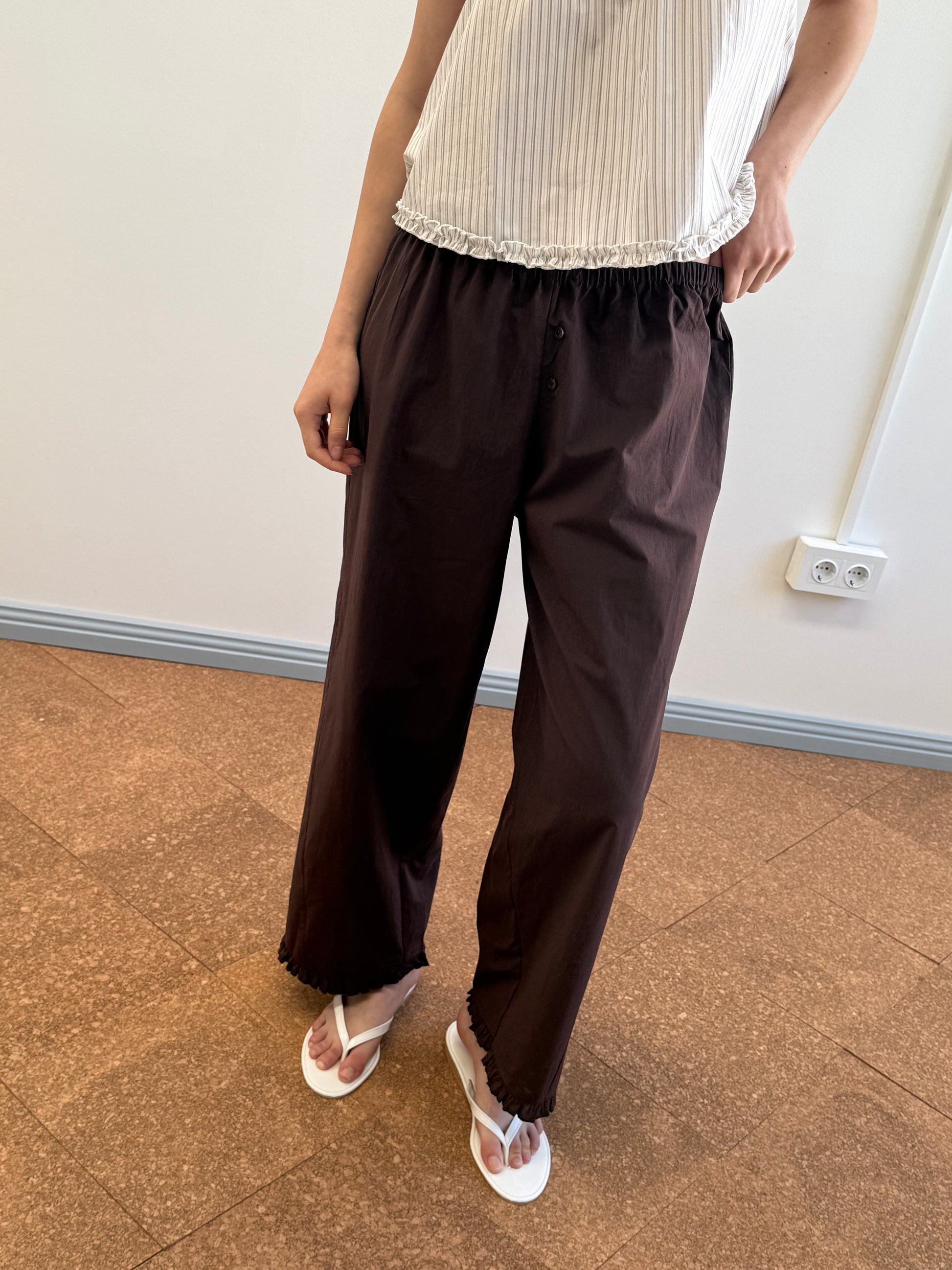 A person stands indoors wearing a sleeveless white top, the Flounce Pant - Umber by Deiji Studios in organic cotton, and white flip-flops. Only the lower torso and legs are visible against a light wall and brown tile floor.
