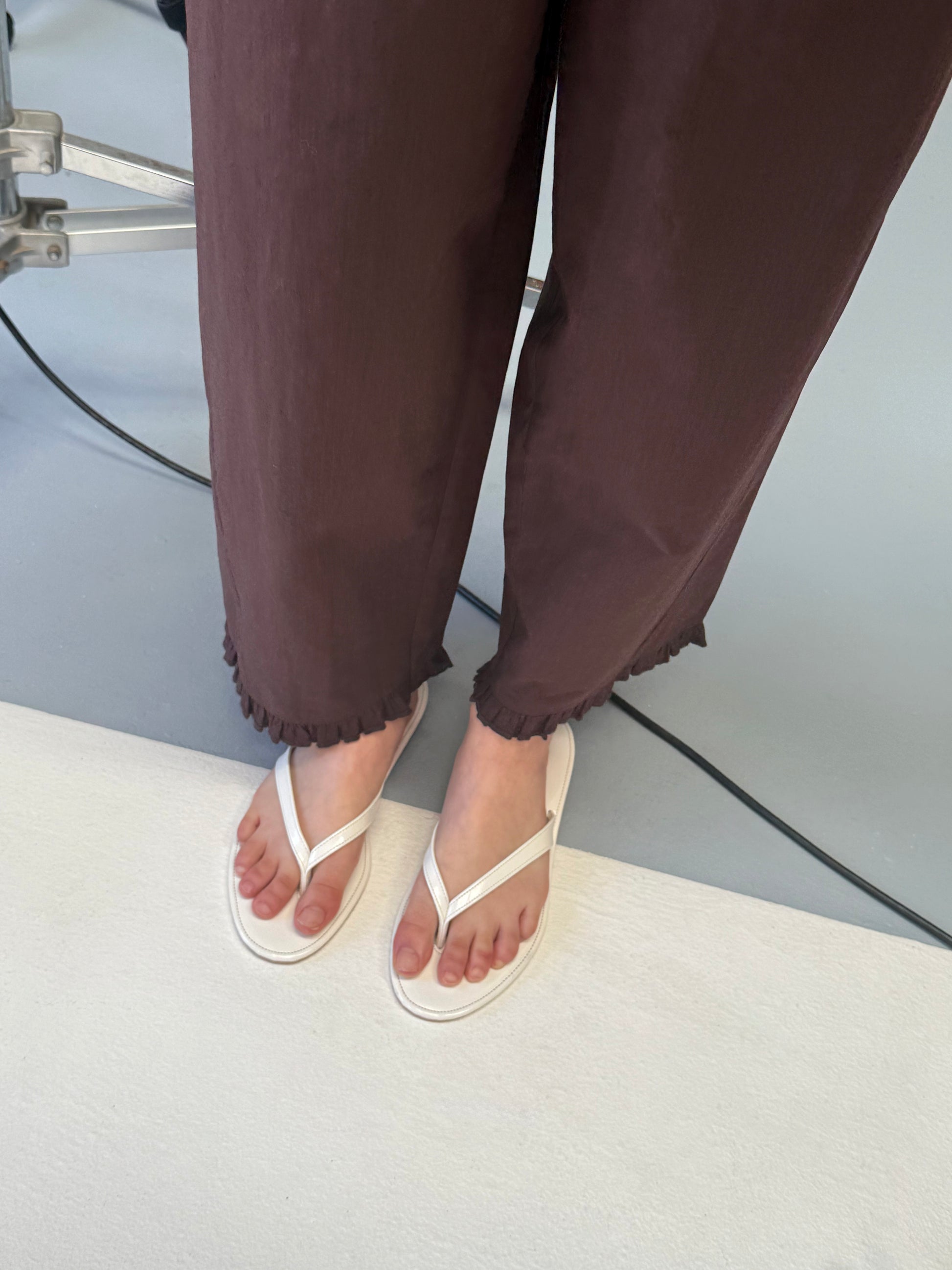 A person in Deiji Studios' Flounce Pant - Umber, featuring ruffled hems, and white flip-flops stands on a light floor beside a black cable and metal equipment.
