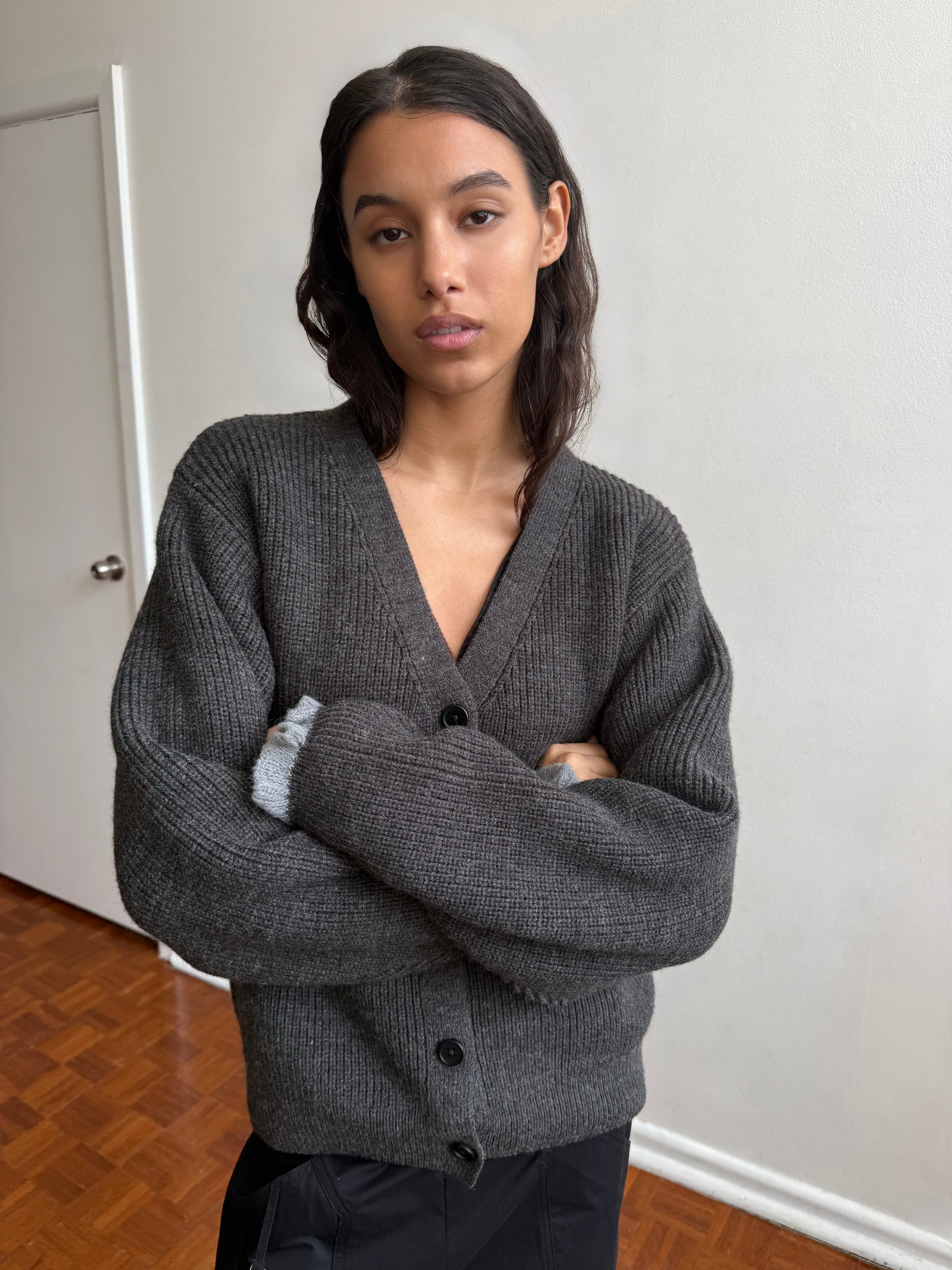 A woman with dark wavy hair stands indoors against a white wall, arms crossed and looking at the camera with a neutral expression. She is wearing the Flounce Cardigan in Charcoal.