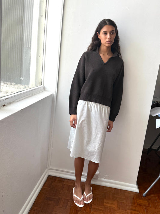 A woman with long dark hair and a neutral expression stands indoors near a window, wearing the Deiji Studios Collared Pullover - Burnt Wood, a white knee-length skirt, and white flip-flops.