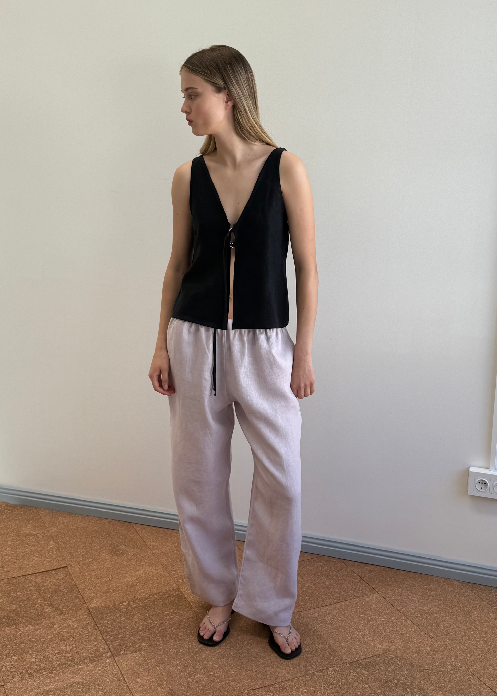 A woman stands indoors against a plain wall wearing the Two Part Top in black with a V-neckline and front tie, paired with loose light beige pants and black sandals for a versatile look. She gazes left, arms relaxed by her sides.