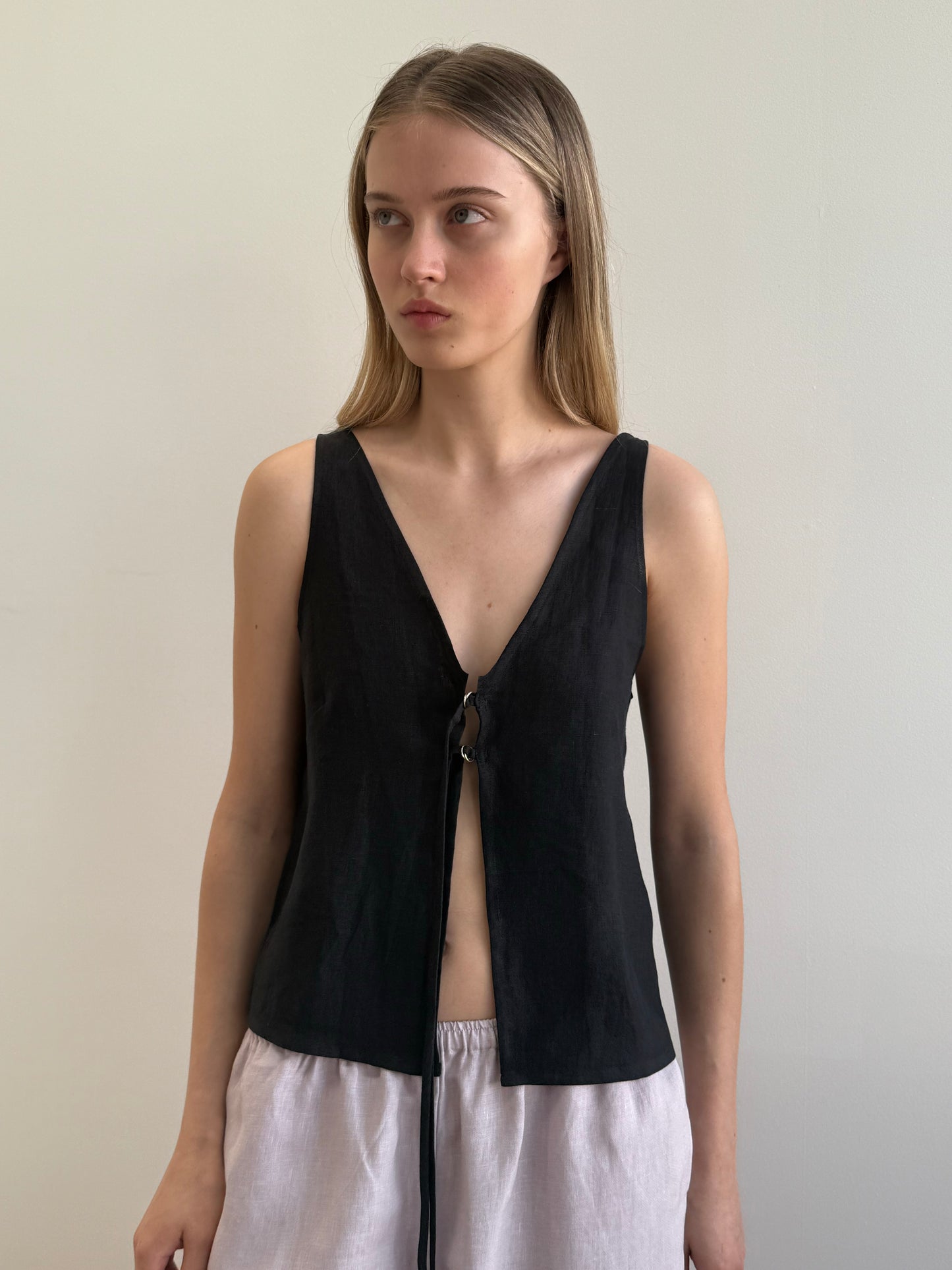 A woman with straight, light brown hair models the Two Part Top - Black, a relaxed top with a deep v-neckline and front tie, paired with light-colored loose pants, against a plain light background.