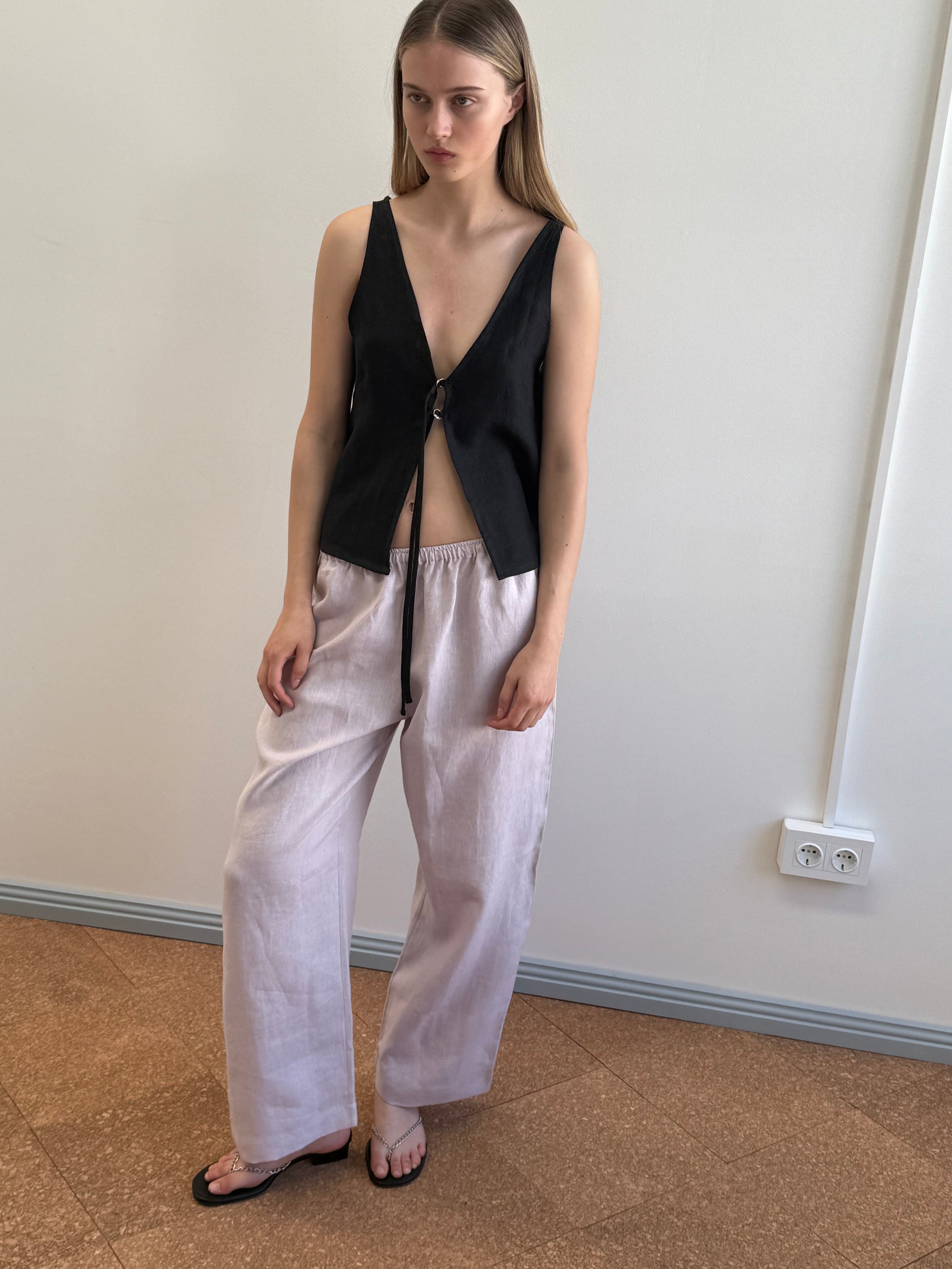 A woman with straight, long hair and a neutral expression stands indoors near a white wall, wearing a sleeveless black deep V-neck top, the Linen Ease Trouser in Lilac, and black sandals.