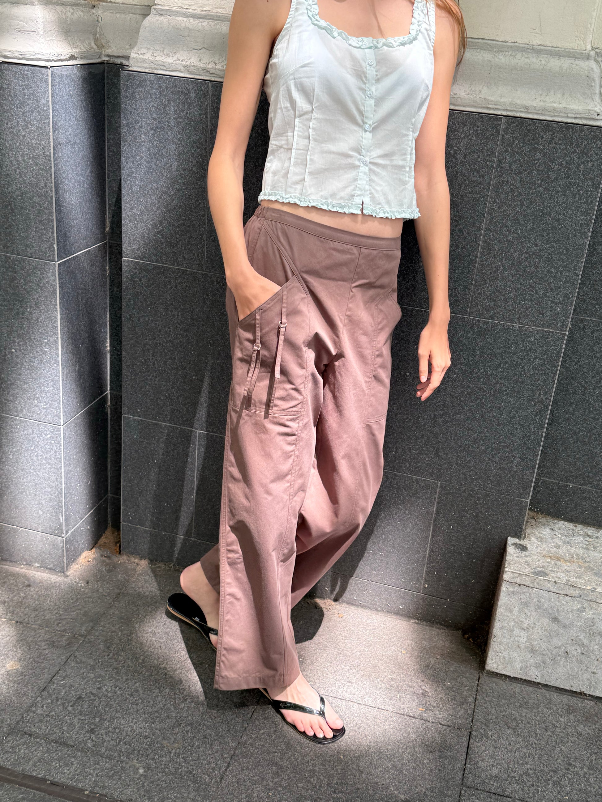 A person in a white sleeveless top, Deiji Studios Adjust Pocket Pant – Sawdust, and black flip-flops stands against a dark tiled wall with one hand in their pocket. Their face isn’t visible.