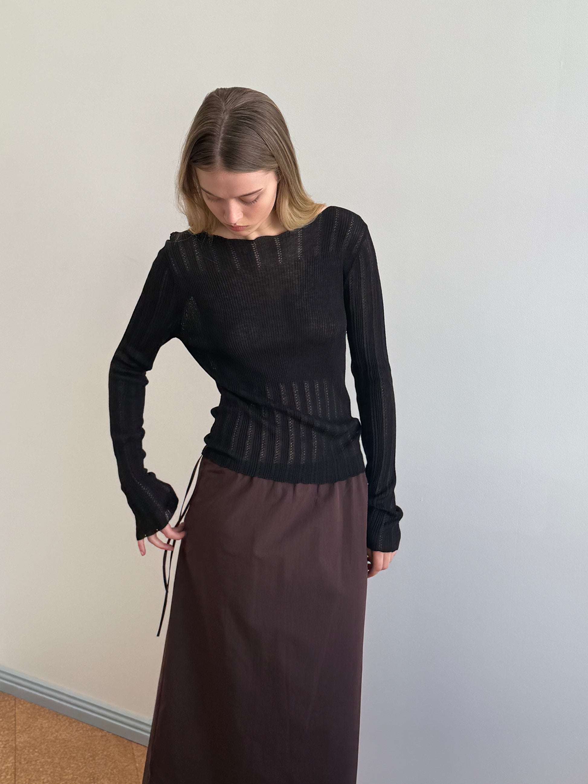 A person with light brown hair looks down, wearing the Deiji Studios Open Work Longsleeve in black and a long brown skirt, standing against a plain light-colored wall.