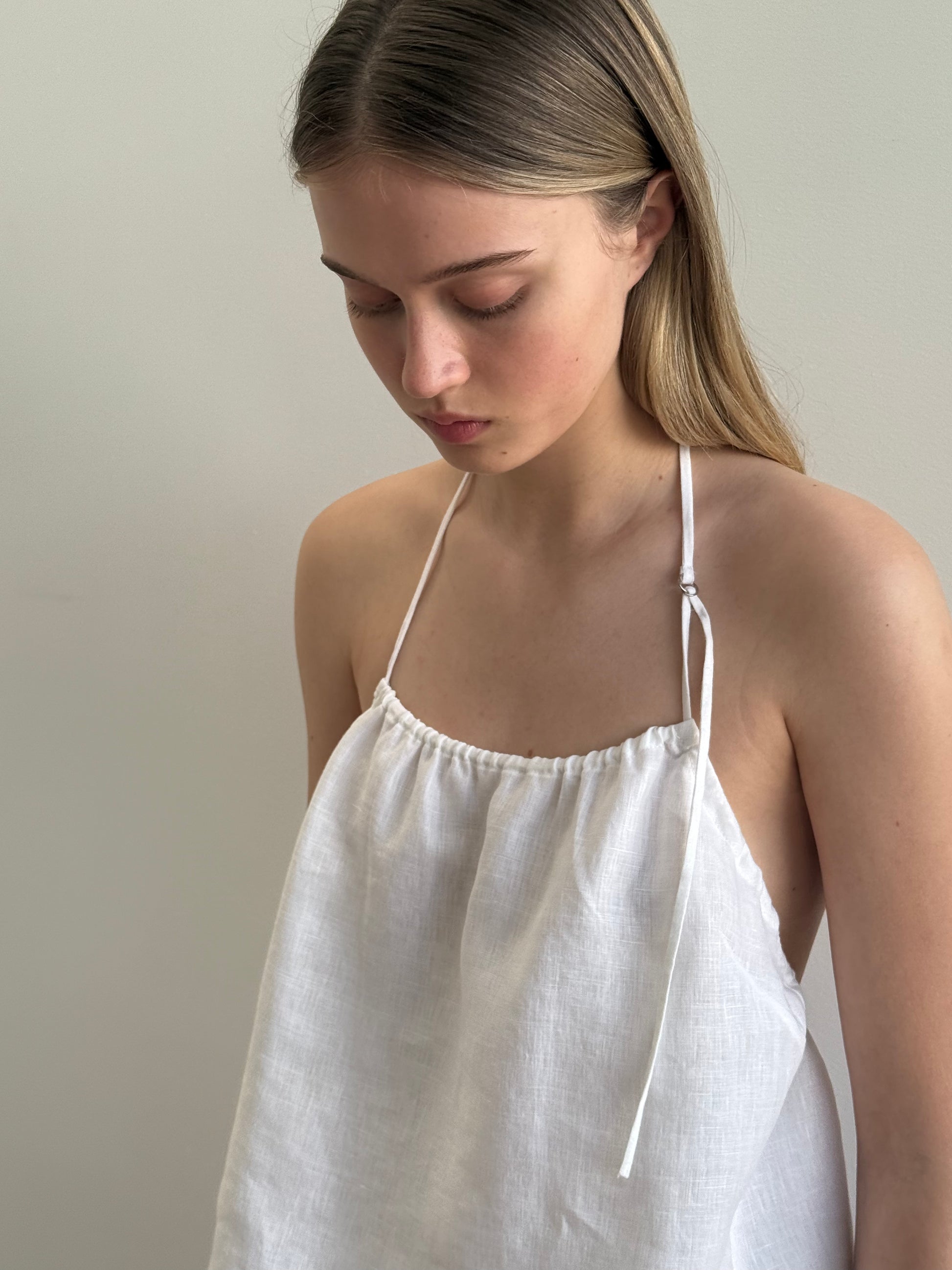 A young woman with long straight hair looks down, wearing the O Ring Halter - White with thin straps. The plain, light background enhances the dress’s soft and minimalistic style.