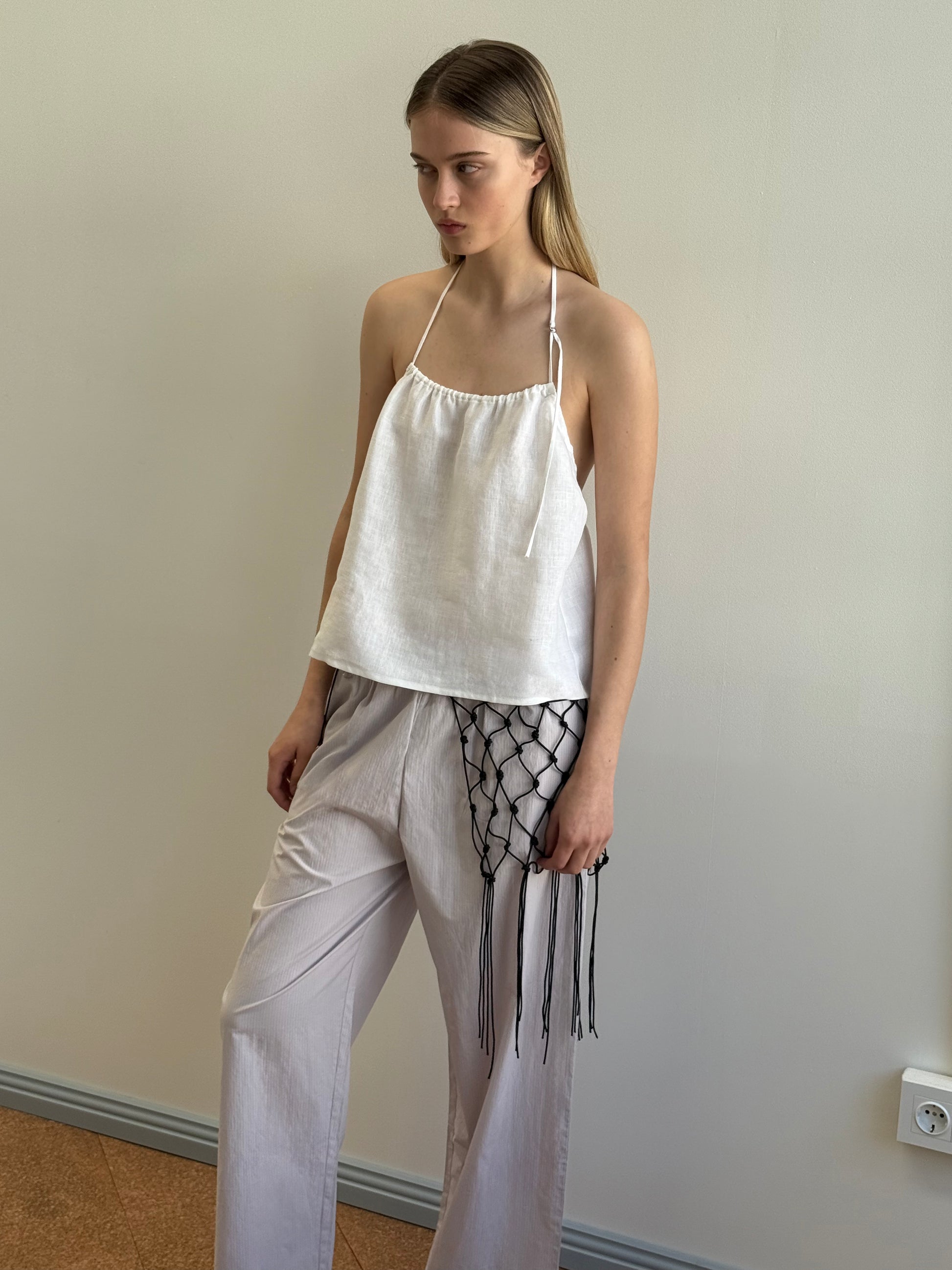 A woman stands against a pale wall, wearing a loose white Deiji Studios halter top and Ease Trouser - Lilac Stripe in organic cotton. She carries a black netted bag with fringe, her expression calm and neutral.