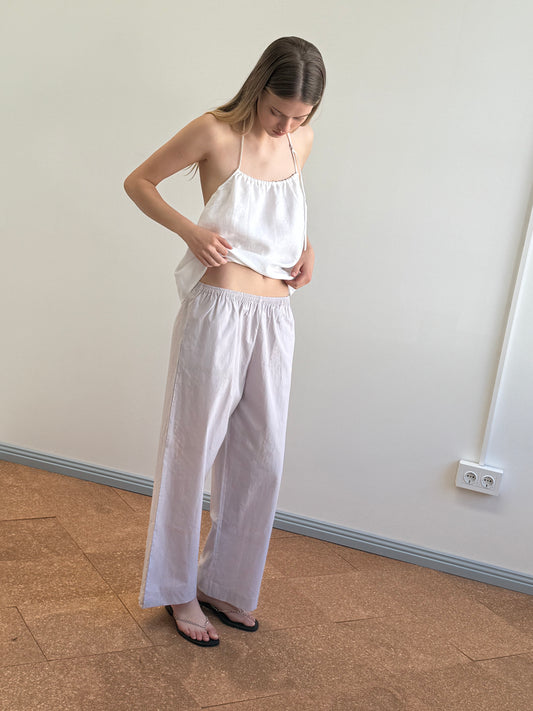 A person with long hair stands indoors on a brown floor, wearing Deiji Studios’ Ease Trouser - Lilac Stripe with an organic cotton sleeveless top, looking down while adjusting their shirt.