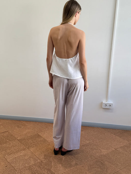 A woman stands indoors facing away from the camera, wearing a white backless halter top and the Ease Trouser - Lilac Stripe by Deiji Studios in light organic cotton with black shoes, near a white wall with an electrical outlet.
